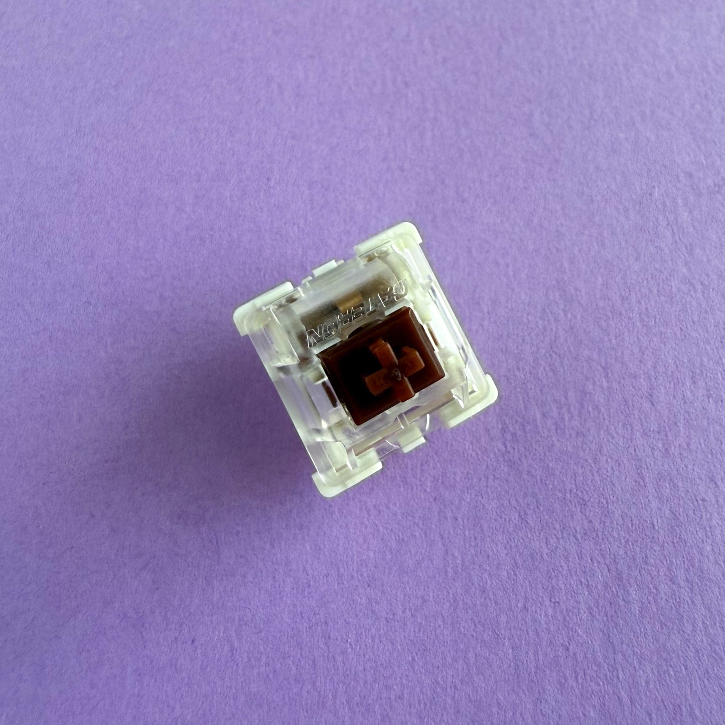 A tactile keyboard switch on a purple background. The switch component is brown, the bottom of the casing is white, and the top of the casing is clear with the word "GATERON" imprinted into it.