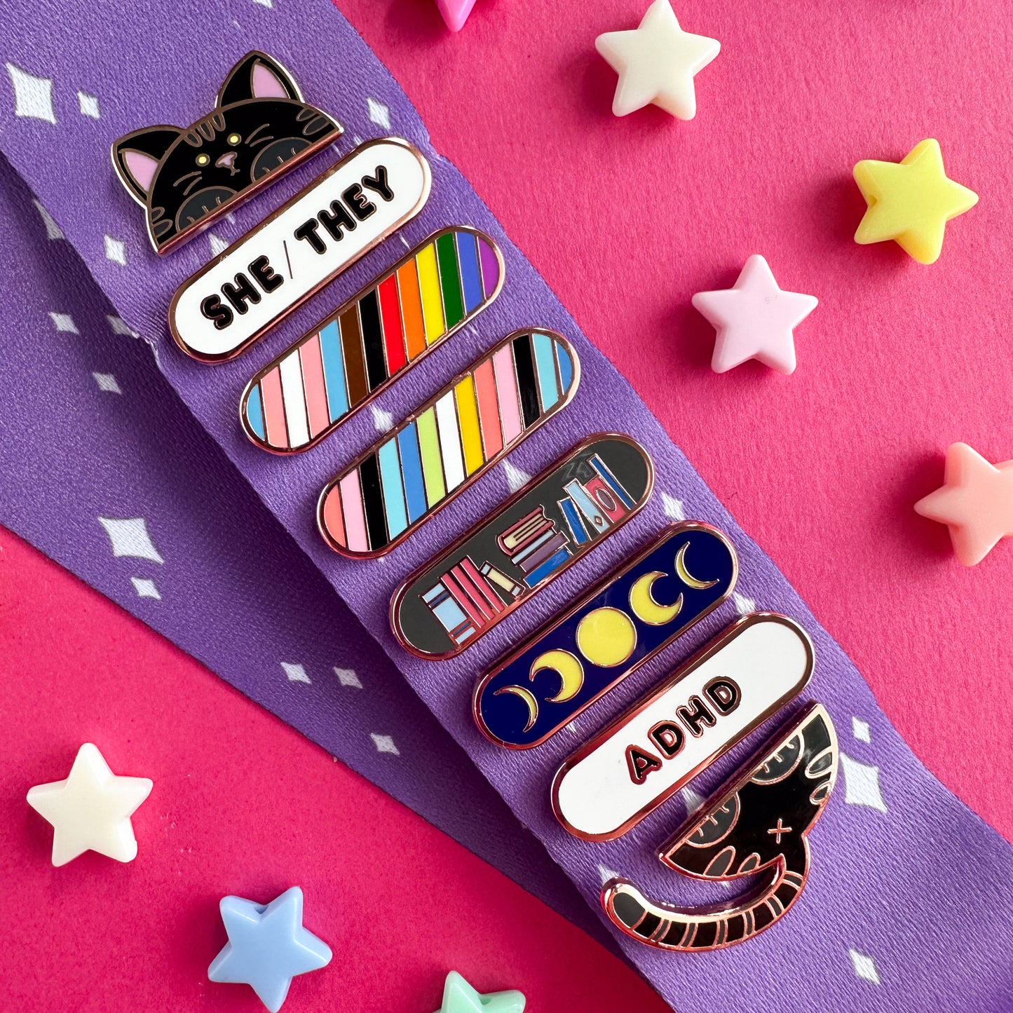 A stack of pins displayed on a purple lanyard with sparkle illustrations on it. The pins are a black cat holding pride flags, books, and pronouns.