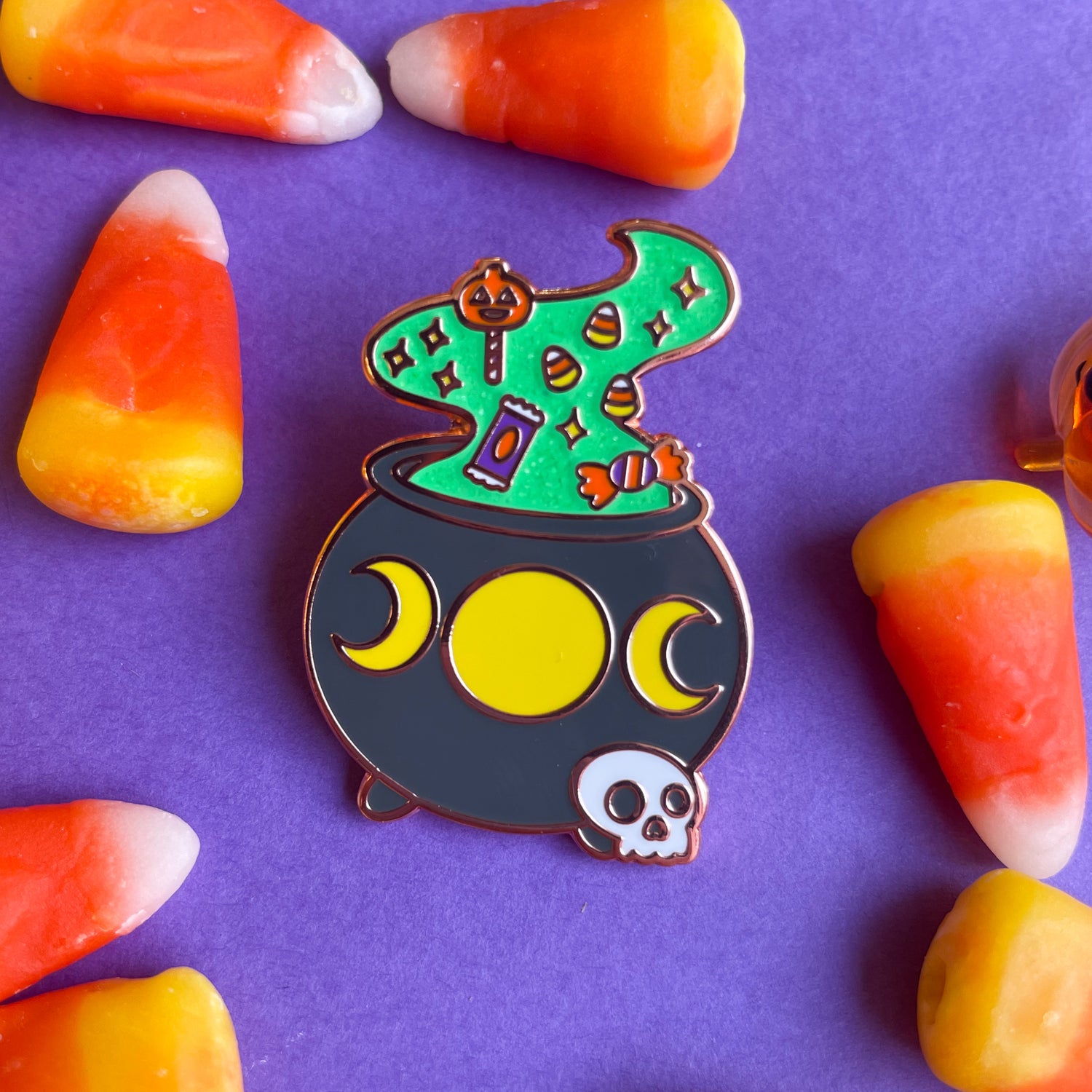 An enamel pin of a cauldron with the trifold moon and magical smoke rising out of it with halloween candy in it. The pin is on a purple background covered in candy corn. 