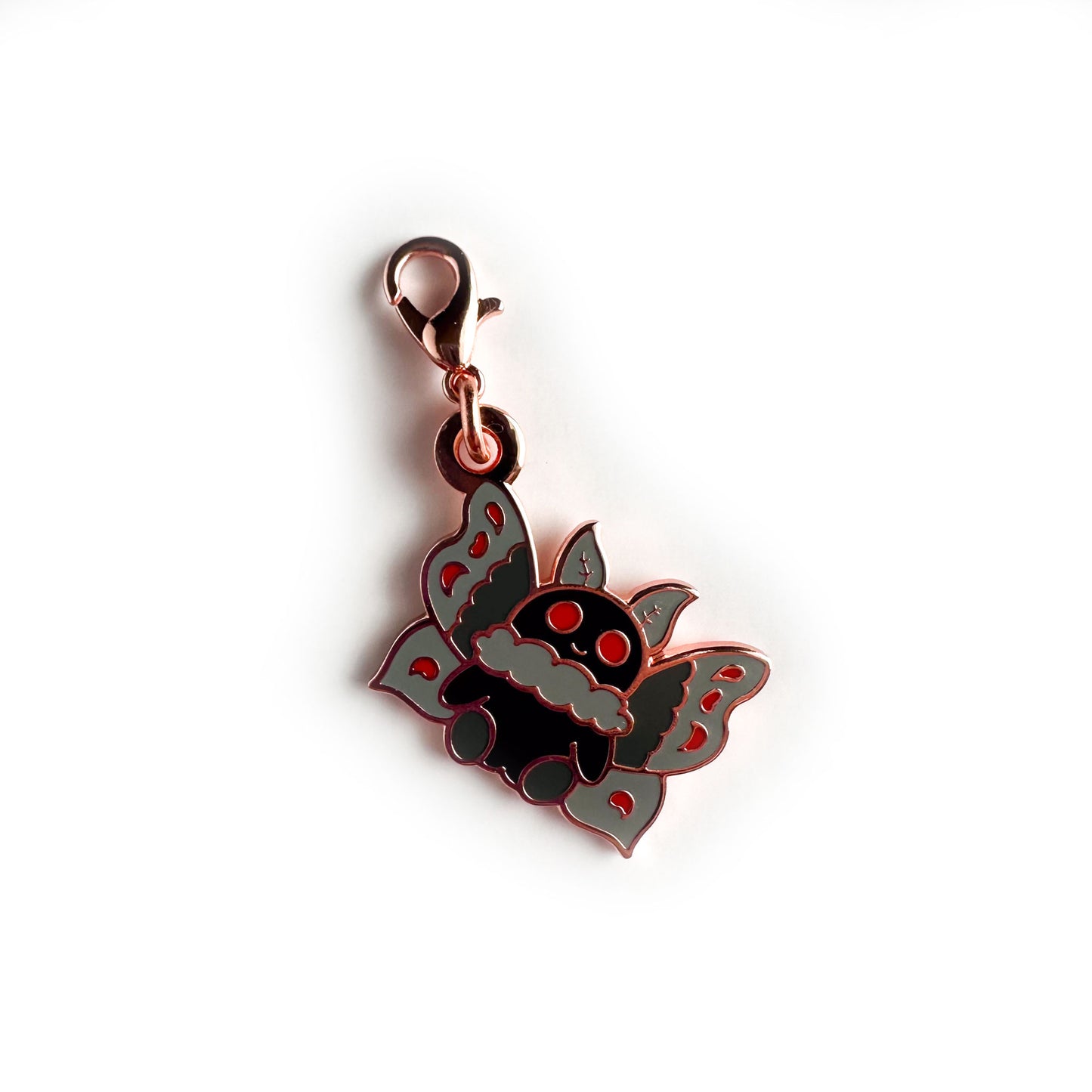 An enamel charm in the shape of a cartoon style mothman with a little smile. The charm has a lobster claw clasp.