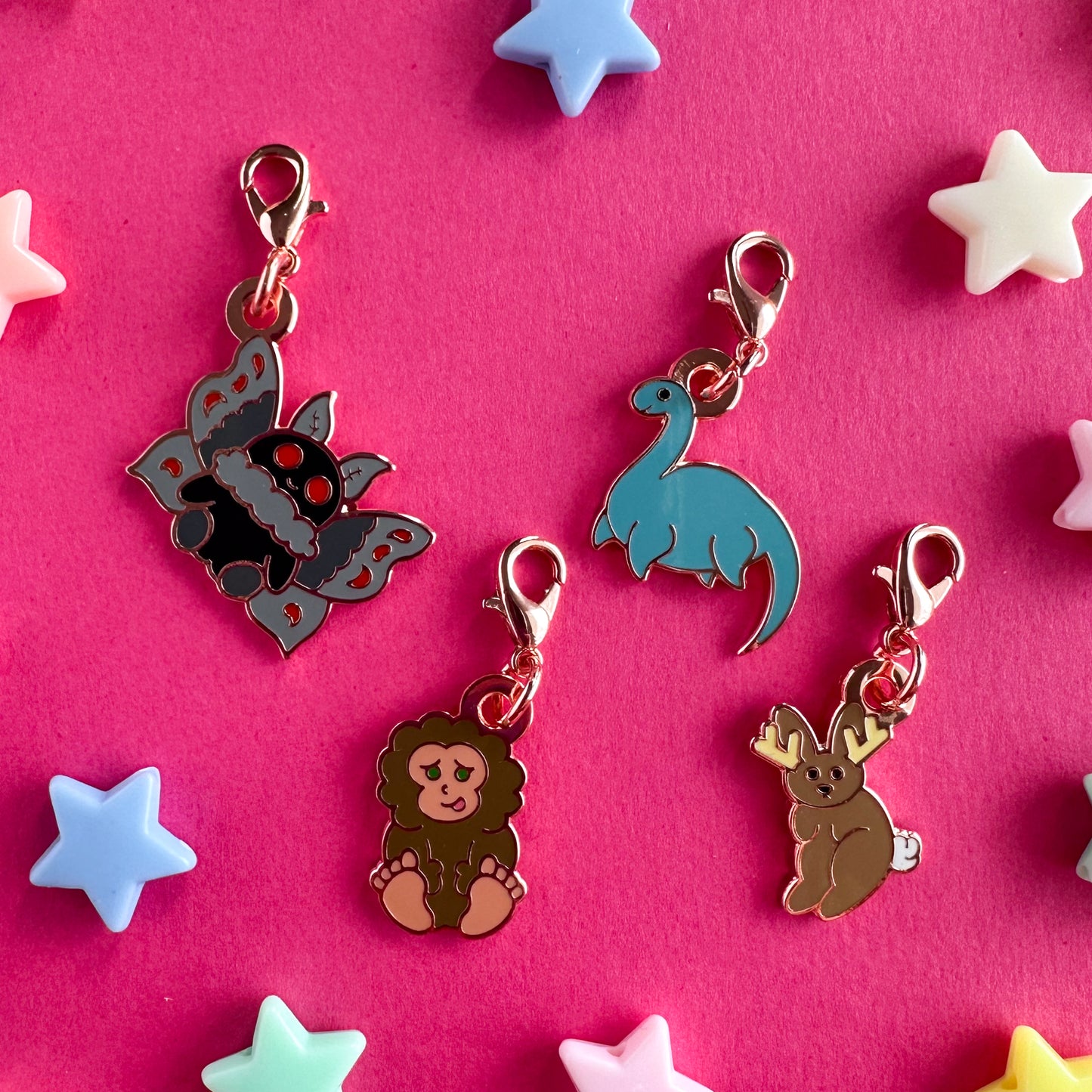 Four charms in the shape of cute cryptids including Mothman, Bigfoot, Nessie, and a jackalope. The charms all have lobster claw clasps. The charms are on a pink paper background with plastic pony beads around them.