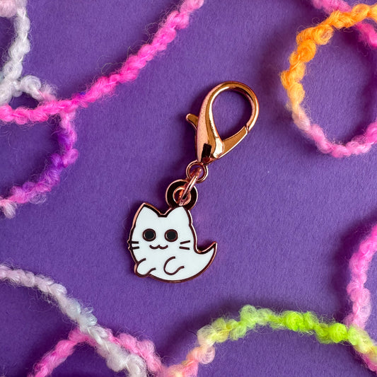 A cute ghost kitty charm with a lobster claw clasp on a purple background surrounded by brightly colored yarn.