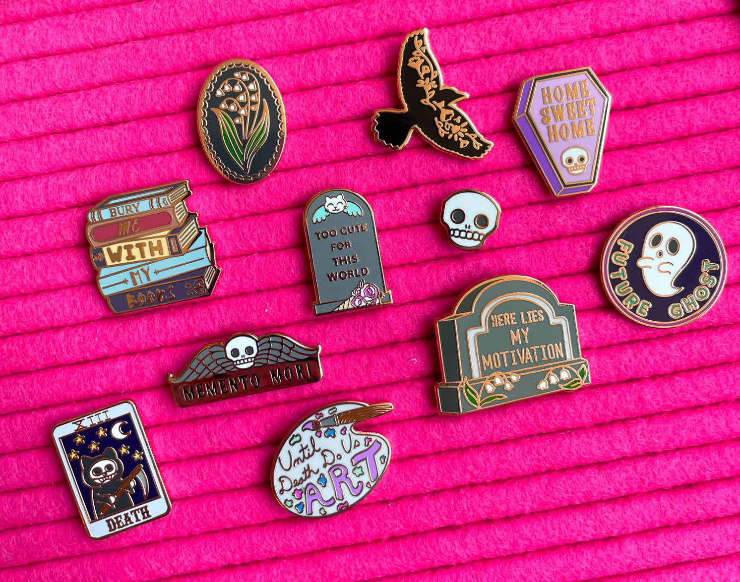 A collection of spooky cute pins on a hot pink background. There is a raven pin, books pin, ghost pin, skull pin, gravestone pins, coffin pin, and artists palette pin.