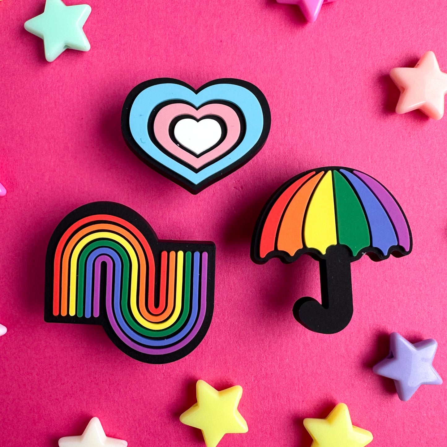 Three PVC charms one in the shape of a heart in the colors of the Trans Pride flag, one a rainbow squiggle, and one a rainbow umbrella. The charms are on a pink paper background with plastic star beads around them.