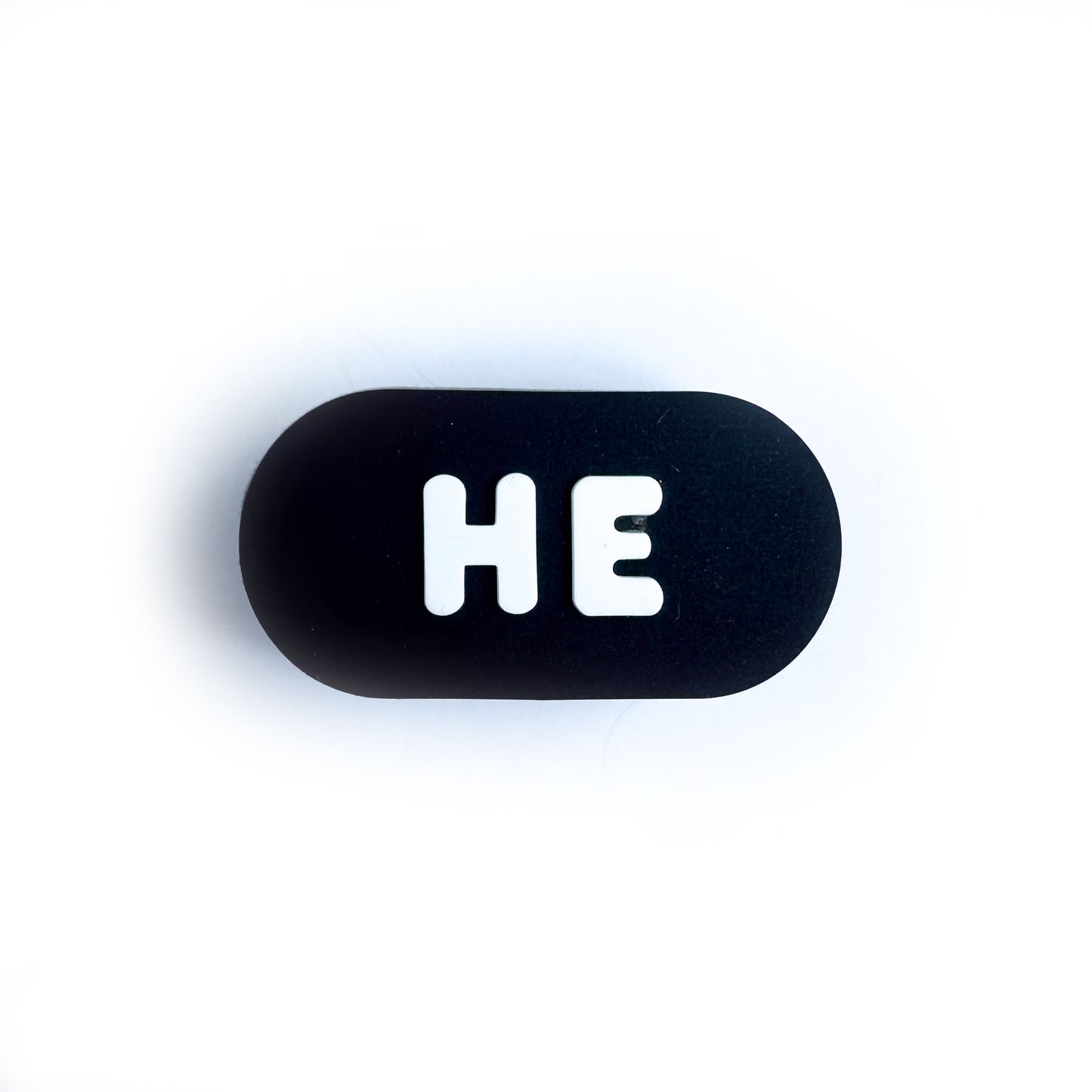 A black oval shaped shoe charm for crocs with white bubble letters that spell out "He"