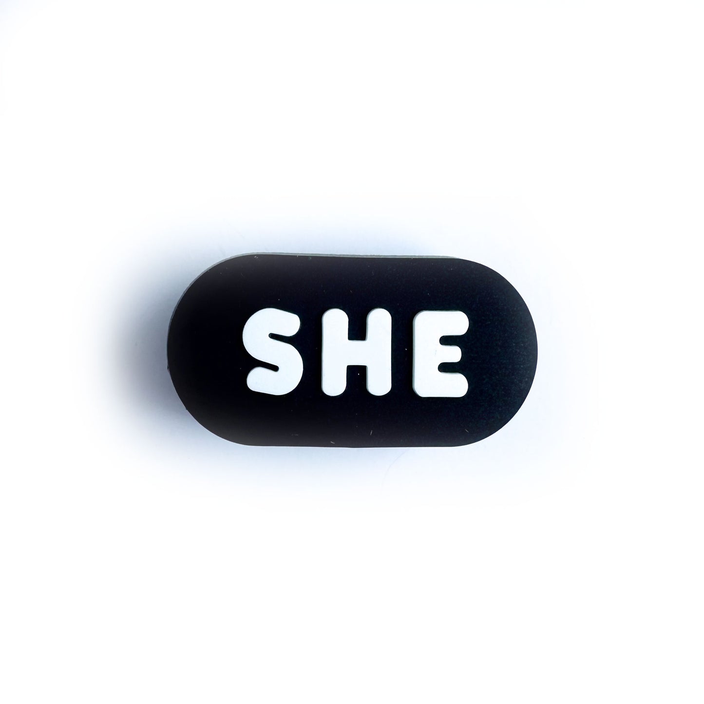 A black oval shaped shoe charm for crocs with white bubble letters that spell out "She"