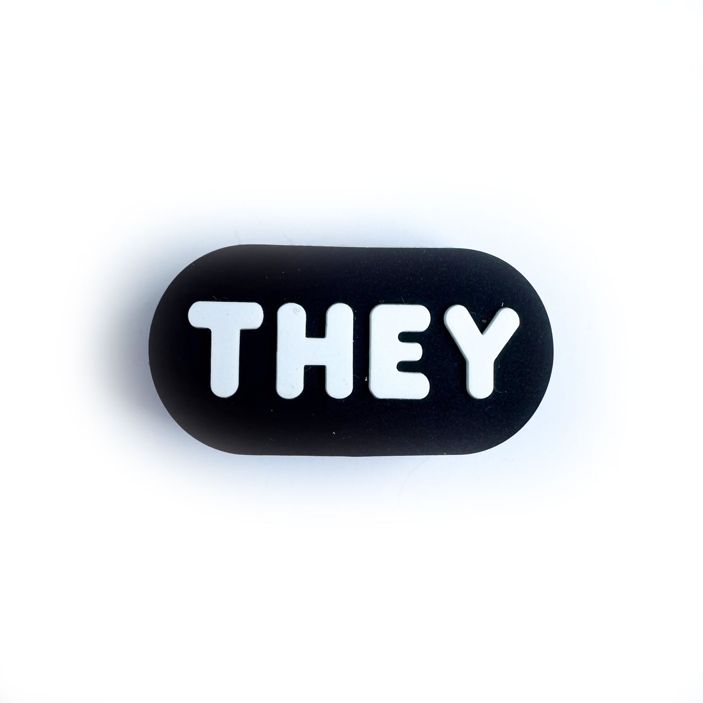 A black oval shaped shoe charm for crocs with white bubble letters that spell out "They"