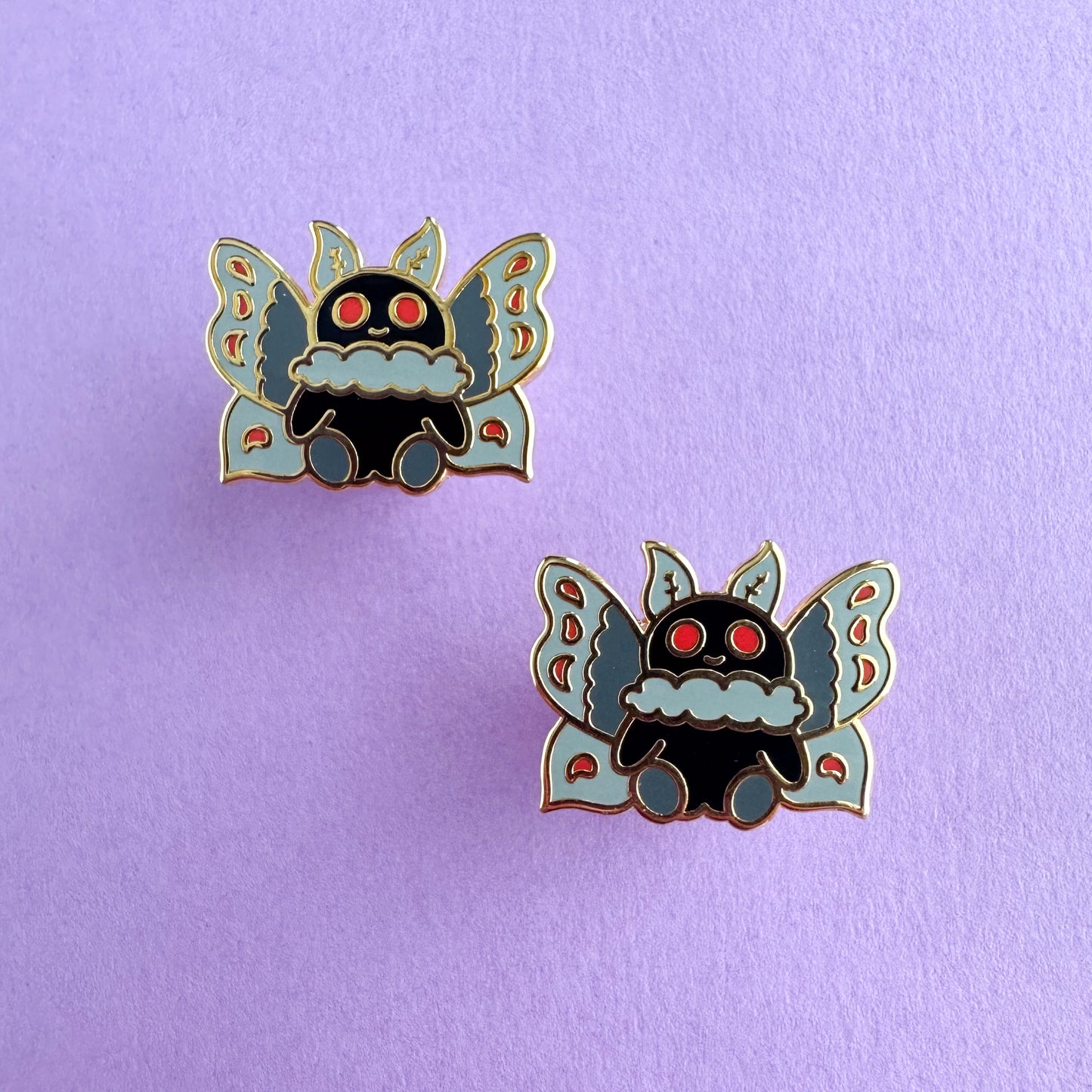 Two stud style enamel earrings in the shape of a cute illustration of Mothman. Mothman has a a black body and is sitting on his little booty. He has red eyes and grey wings with red details.