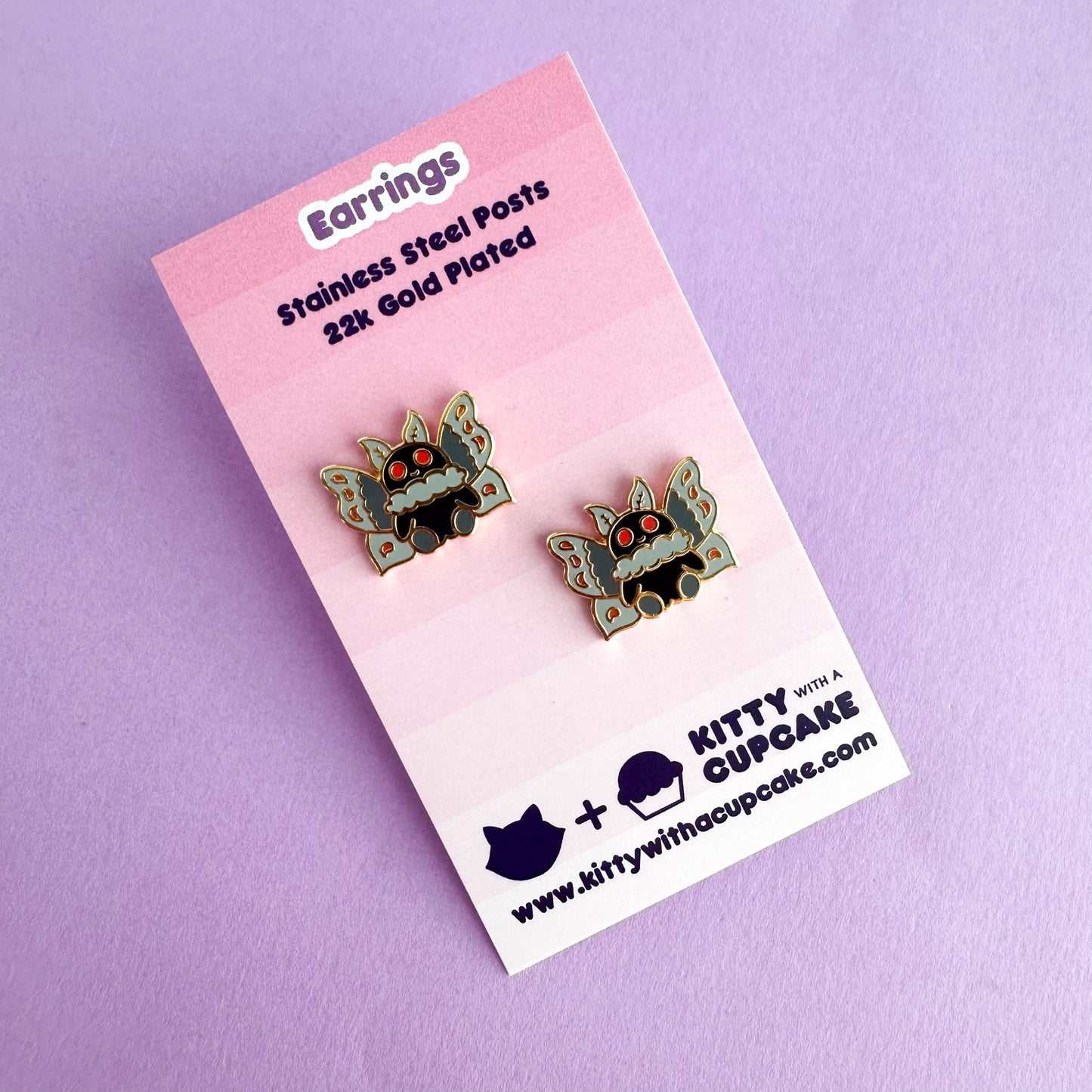 Two stud earrings in the shape of Mothman packaged on a pink card that reads "Earrings Stainless Steel Posts 22k Gold Plated"