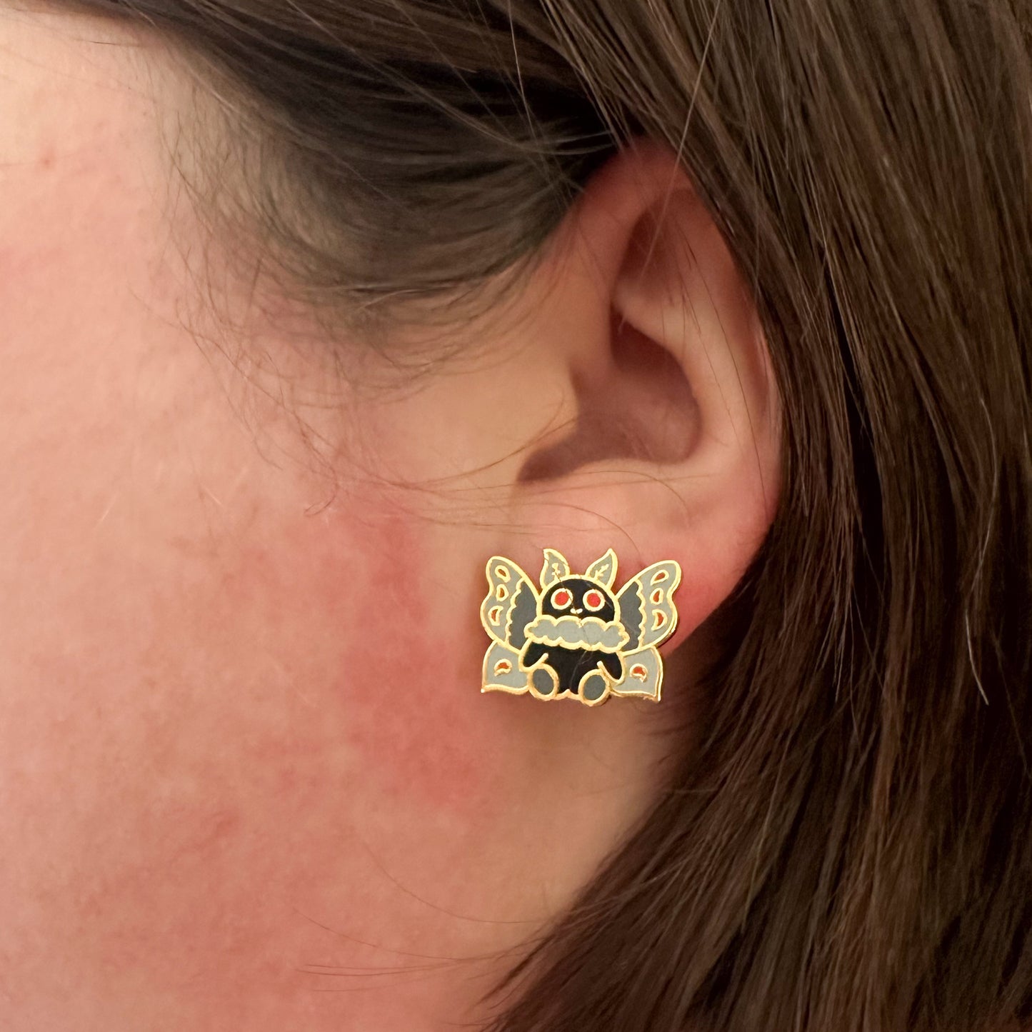 A close up of a white woman's ear with brown hair. There is a stud earring in the shape of Mothman on her ear.