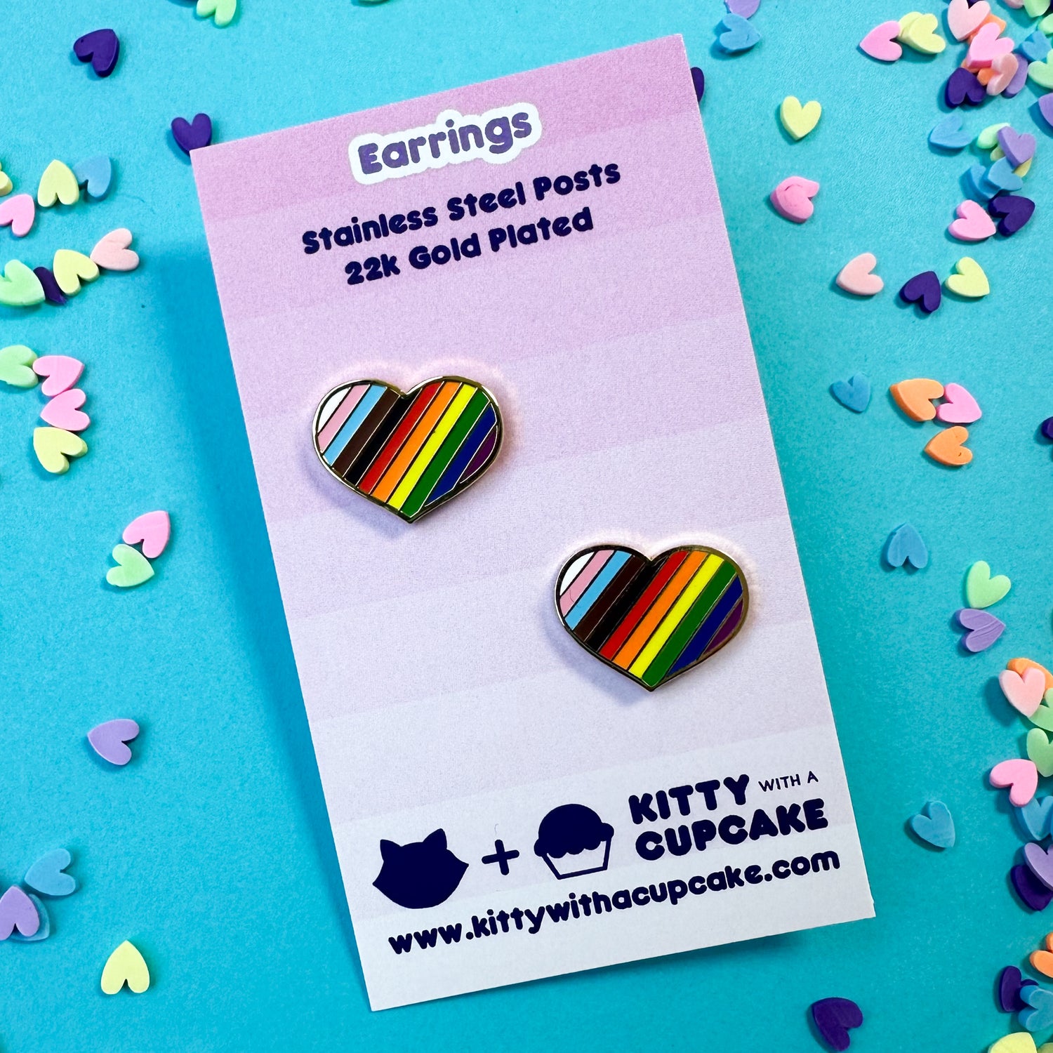 Two chunky heart earrings with diagonal stripes on them in the progress pride flag colors. The earrings are packaged on a pink card that is sitting on top of a blue background with confetti hearts around it.
