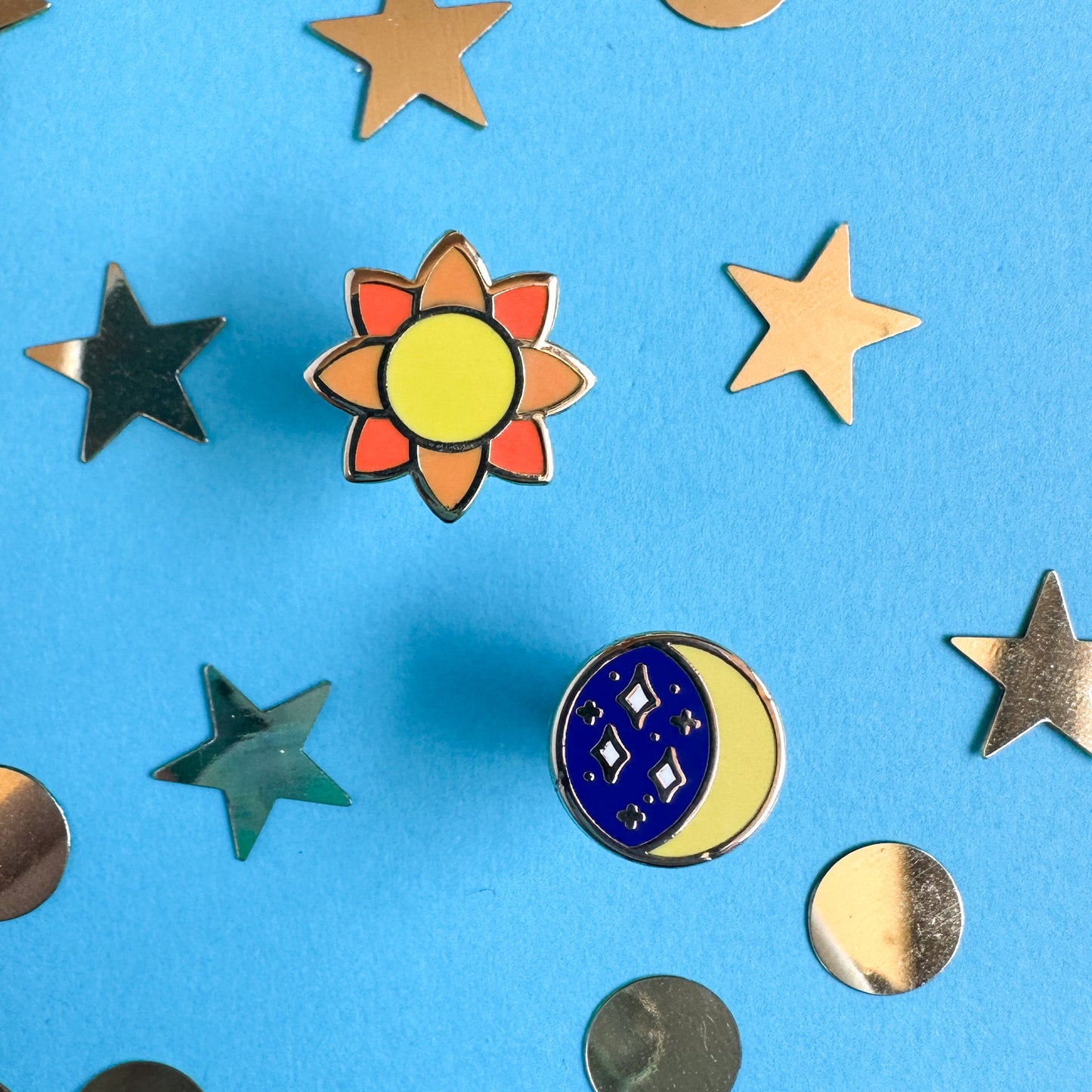 A set of earrings on a light blue background with silver star confetti. One earring is sun shaped with a yellow circle center, and petals of two shades of orange. The other is a circle, with a yellow crescent moon and the rest of the circle is blue with stars.