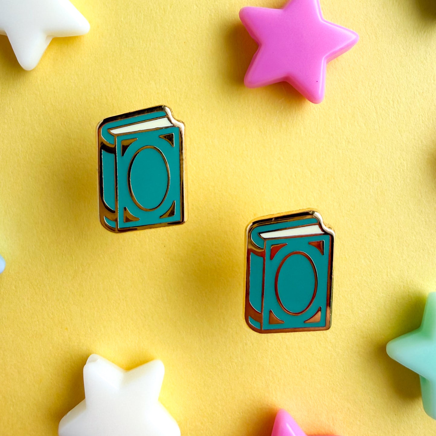 A pair of teal book shaped stud earrings with plastic star beads around them on a yellow background.