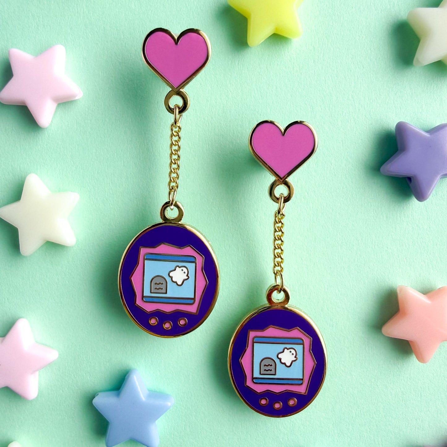 Pair of earrings with hearts connected to a virtual pet shaped charm with a ghost illustration on the screen on a mint green background with plastic star beads around.