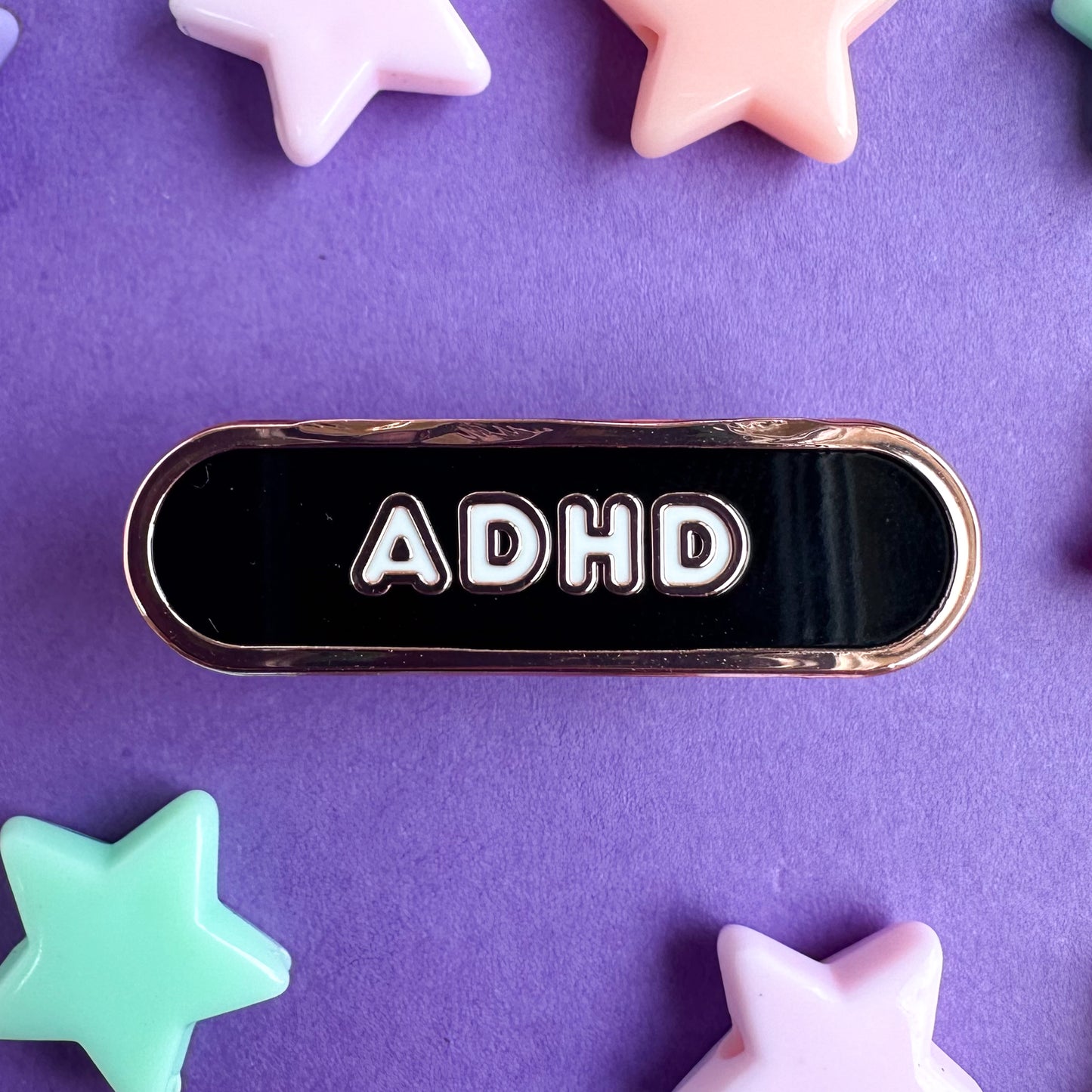 A black oval shaped enamel pin with the letters ADHD on it in white bubble letters. The pin is on a purple paper background with pastel star beads strewn around it.