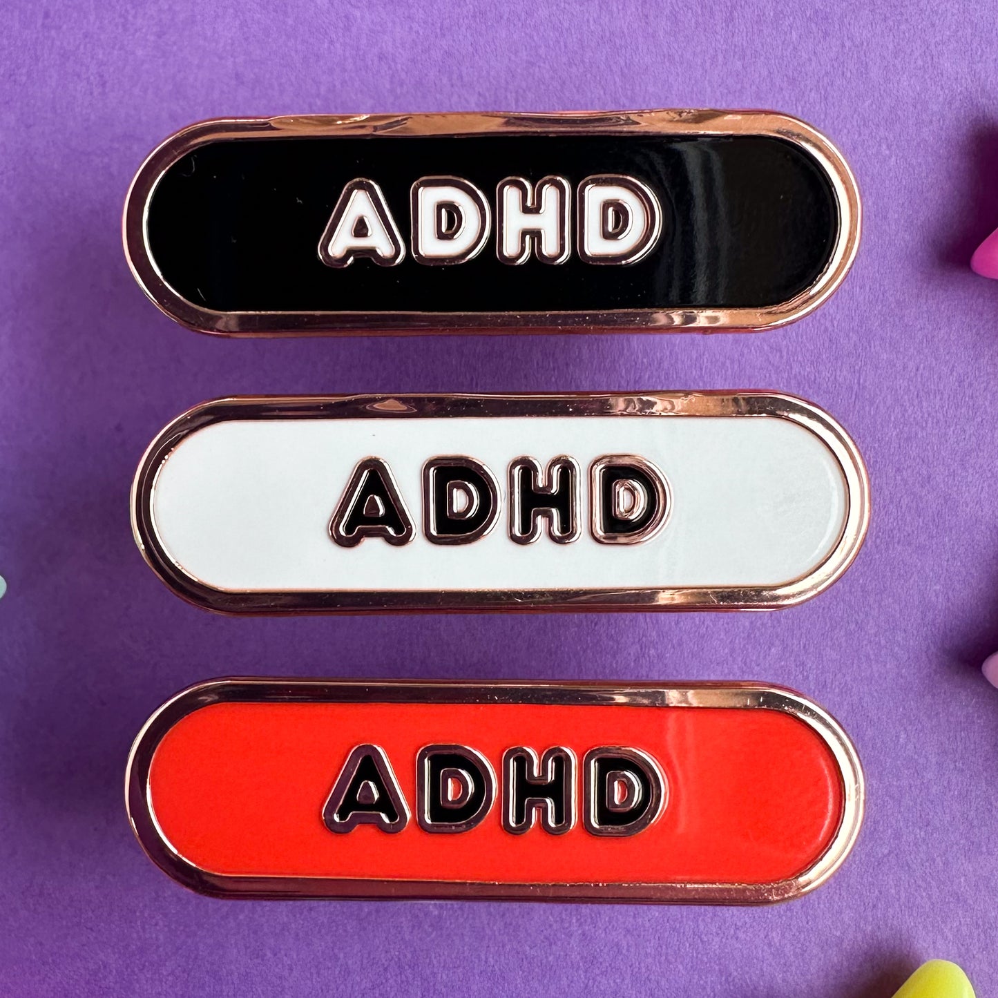 Three capsule shaped pins that have the letters ADHD on them, the top is black, the middle is white, and the bottom is red. The pins are on a purple paper background.