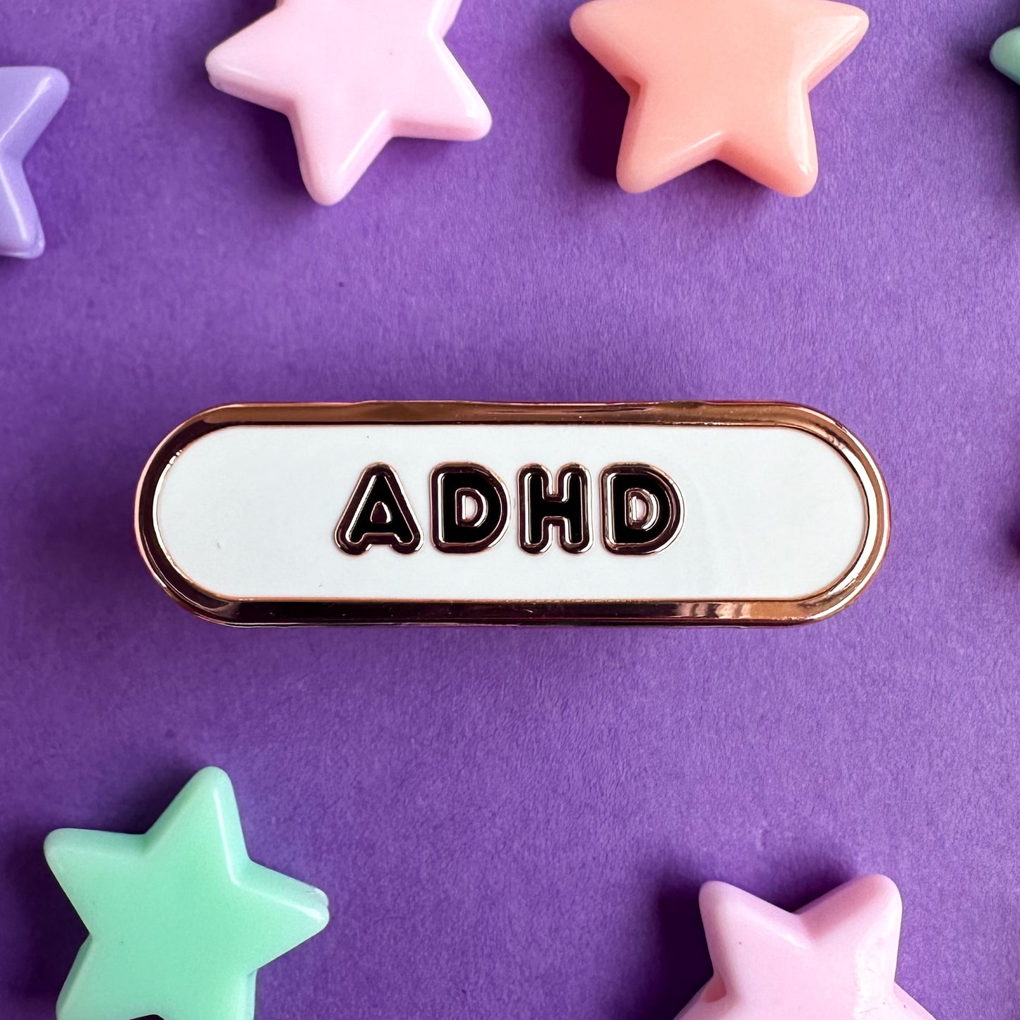 An enamel pin in the shape of a capsule with a white background and black bubble letters that read ADHD. The pin is on a purple background with plastic star beads scattered around it.