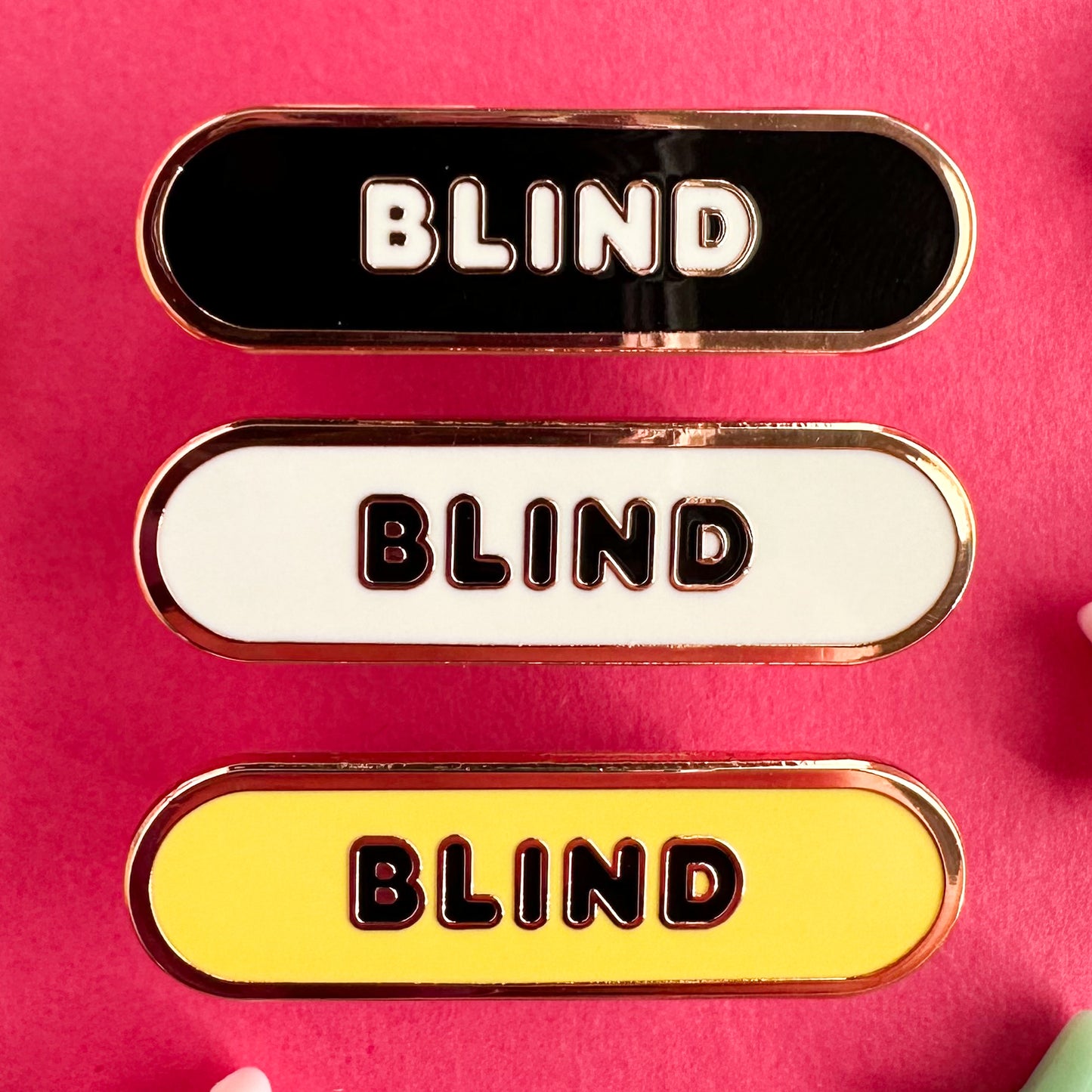 Three enamel pins in the shape of a capsule that all have bubble letters that read "Blind" the top pin has a black background, the middle is white, the bottom is yellow. The pins are on a pink paper backdrop.