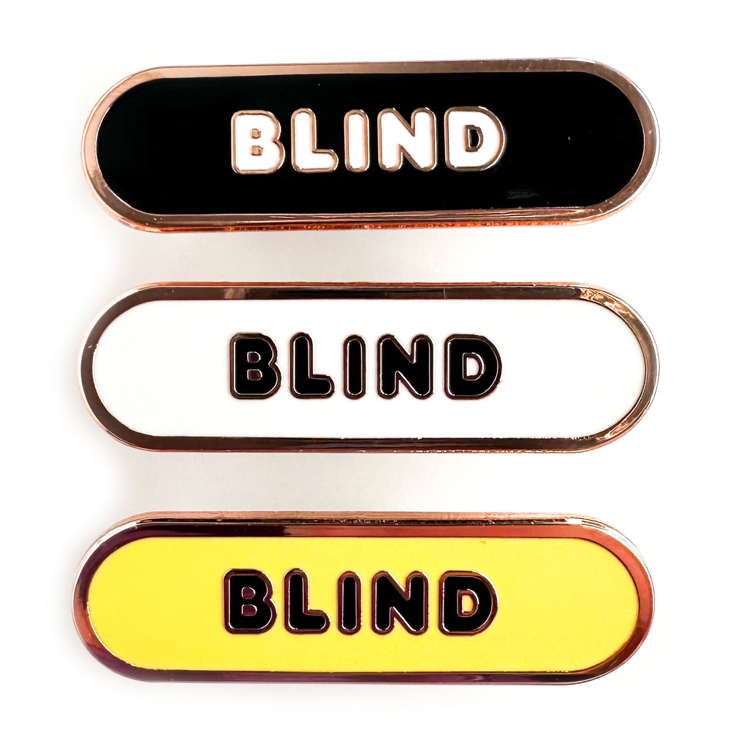 Three enamel pins in the shape of a capsule that all have bubble letters that read "Blind" the top pin has a black background, the middle is white, the bottom is yellow.
