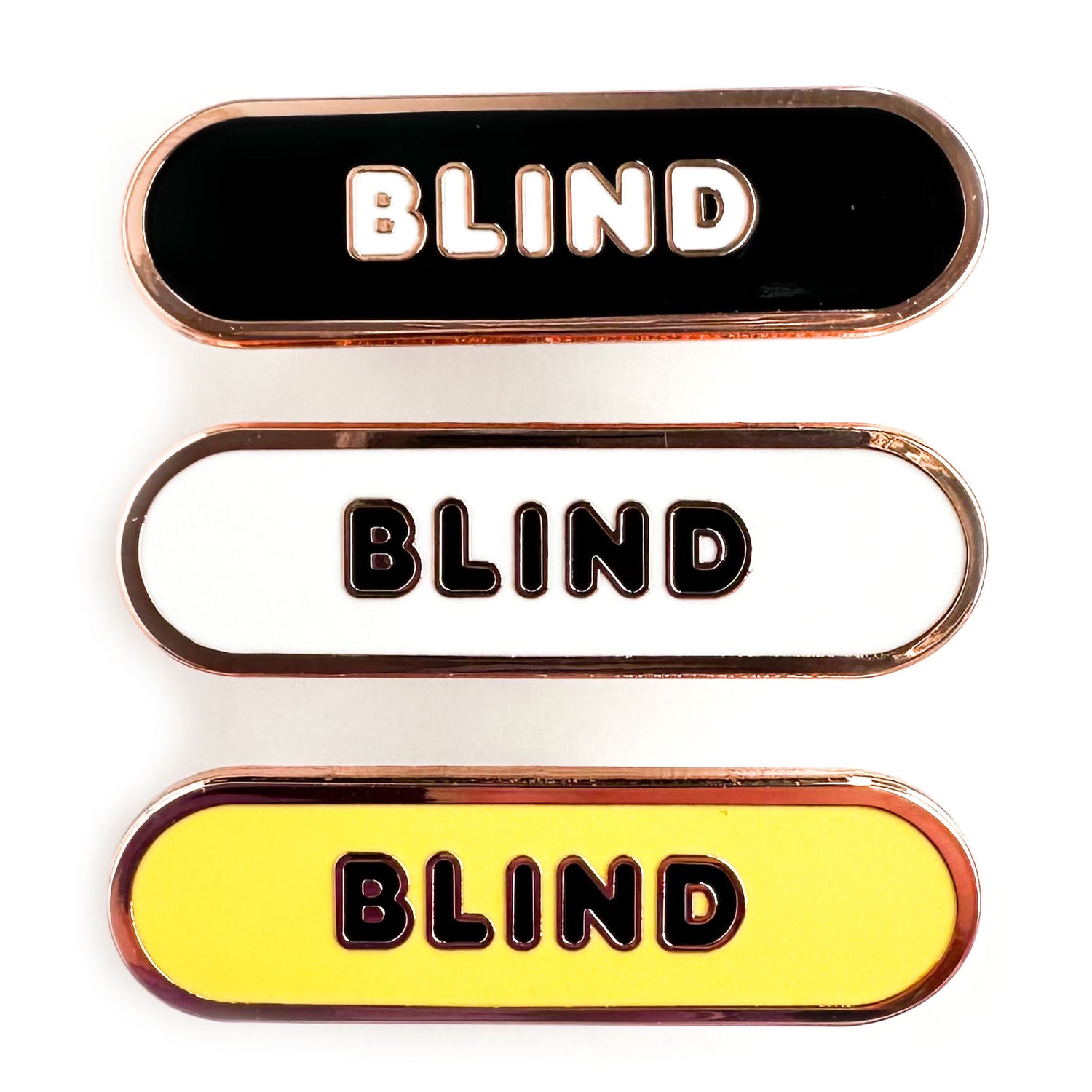 Three enamel pins in the shape of a capsule that all have bubble letters that read "Blind" the top pin has a black background, the middle is white, the bottom is yellow.