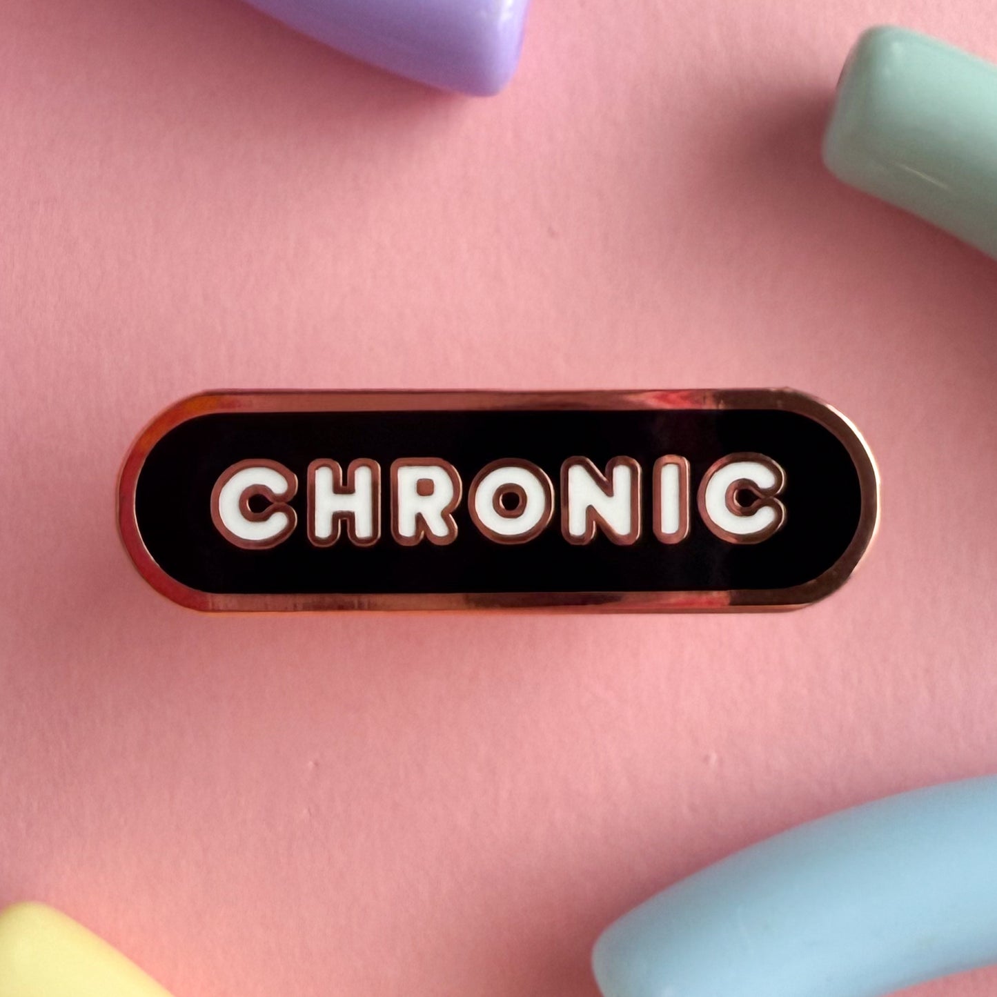 A bandaid shaped pin with the text CHRONIC in bubble letters. The background is black and the text is white. The pin is on a pink background.