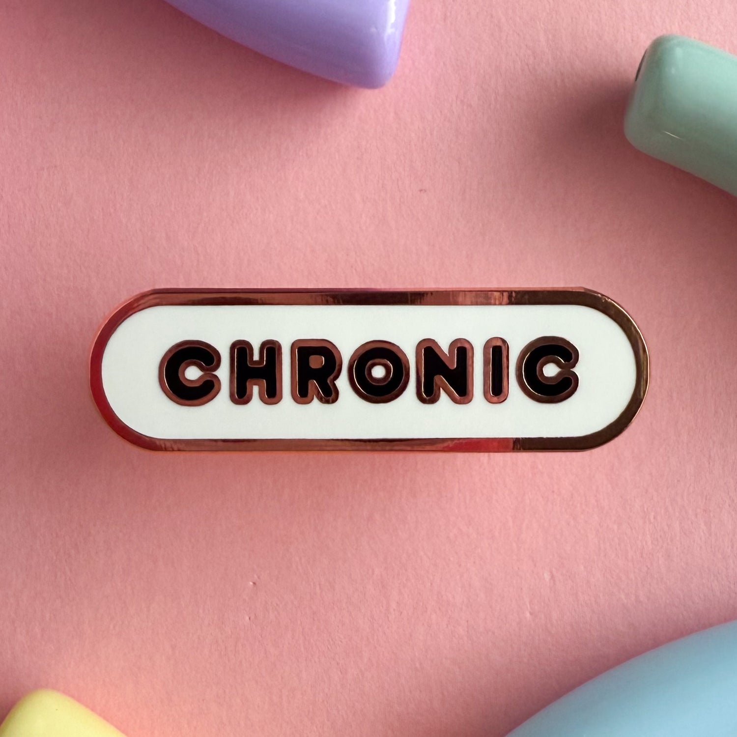A bandaid shaped pin with the text CHRONIC in bubble letters. The background is white and the text is black. The pin is on a pink background.
