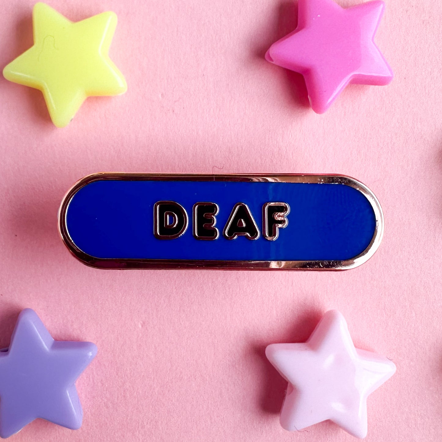 A blue capsule shaped pin with black bubble letters that spell "Deaf". The pin is on a pink background with plastic star beads around it.