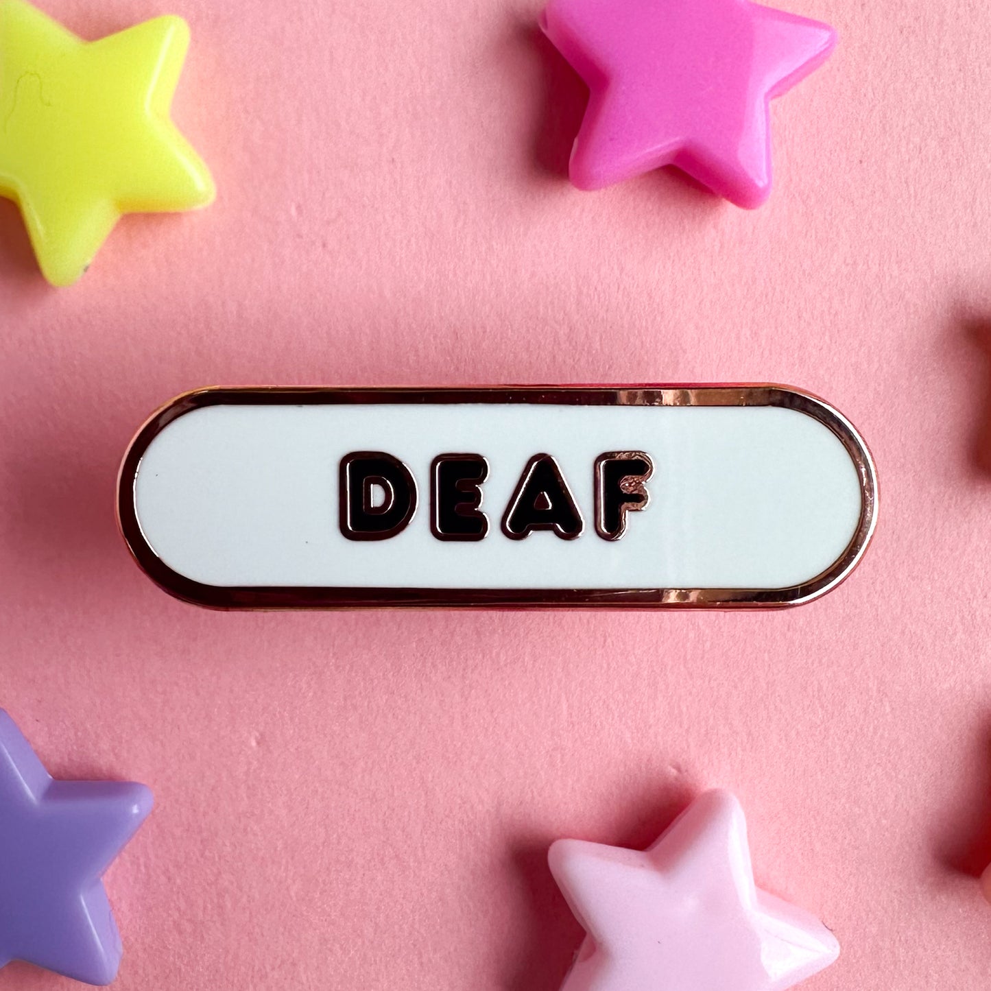 A white capsule pin with black bubble letters. The pin is on a pink background with star beads around it.