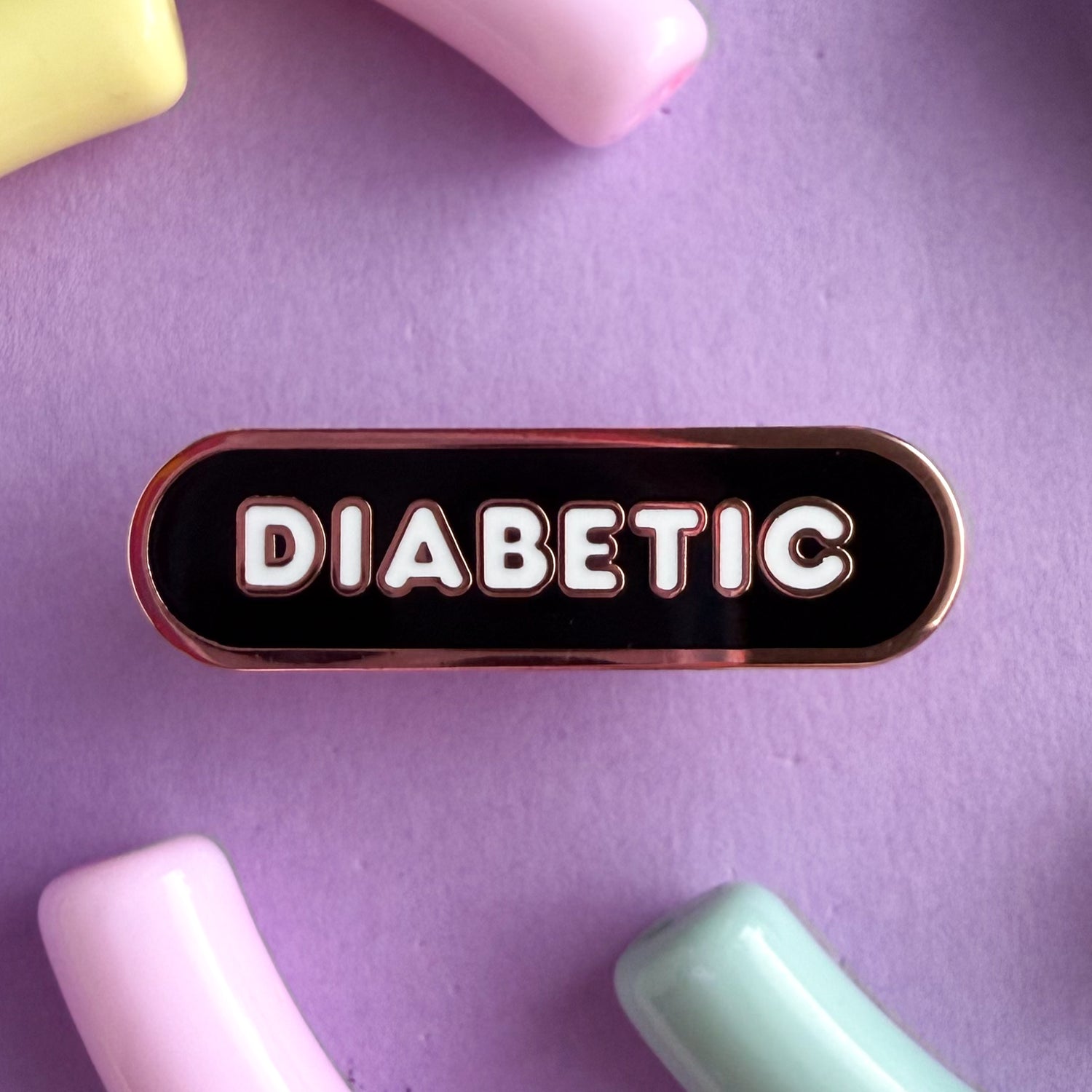 A bandaid shaped pin with the text DIABETIC in bubble letters. The background is black and the text is white. The pin is on a purple background.