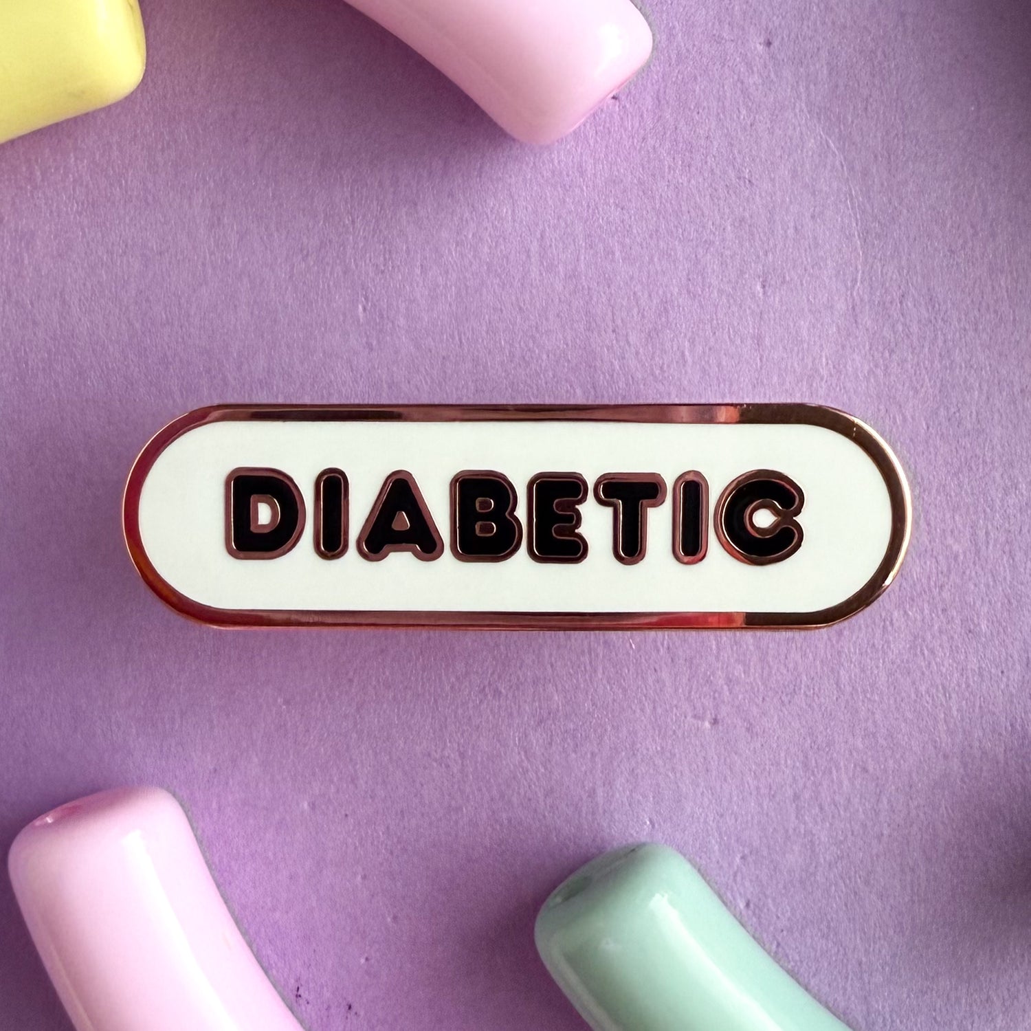 A bandaid shaped pin with the text DIABETIC in bubble letters. The background is white and the text is black. The pin is on a purple background.