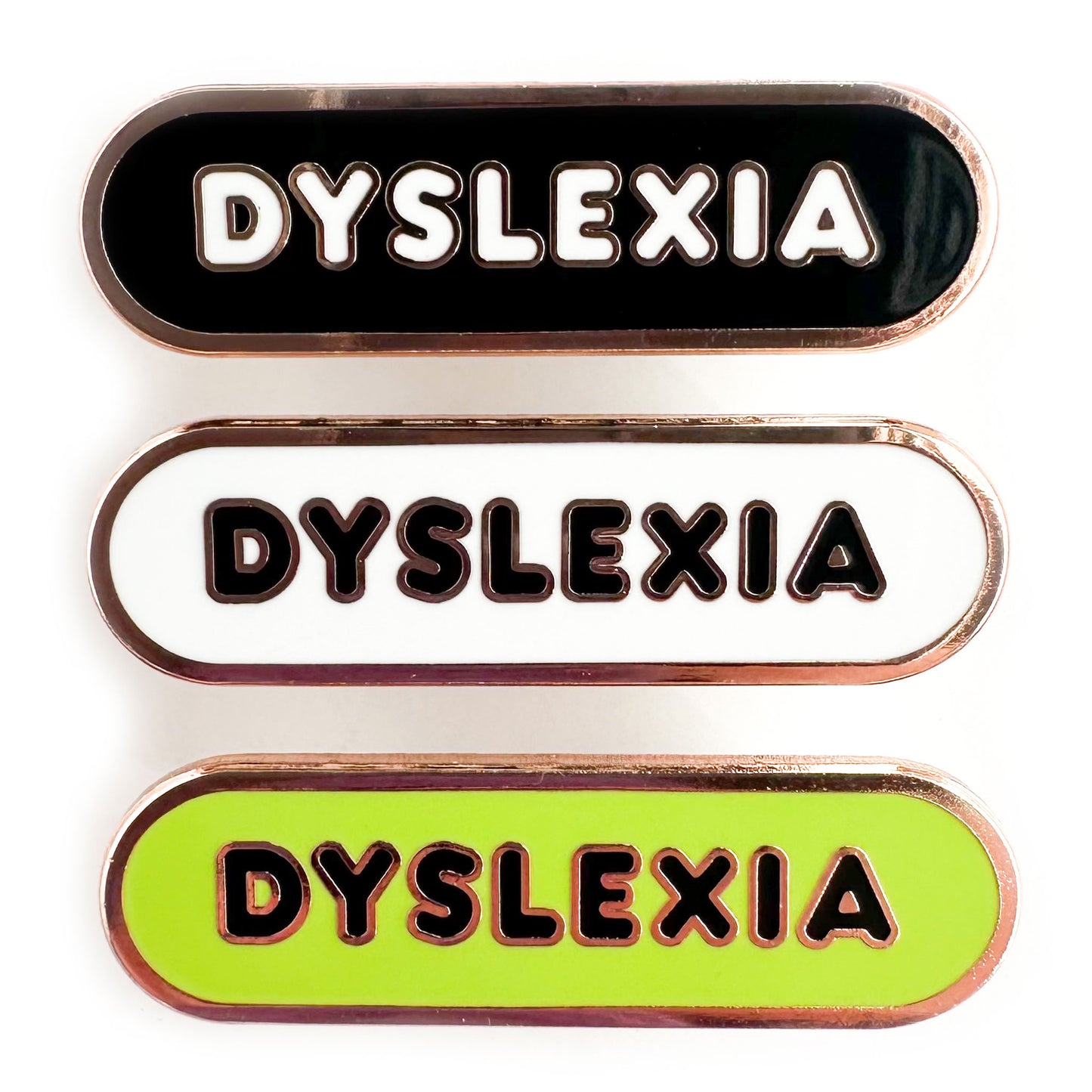 Three enamel pins in a stack that all read "Dyslexia" in bubble letters. The top pin is black, the middle is white and the bottom is lime green.