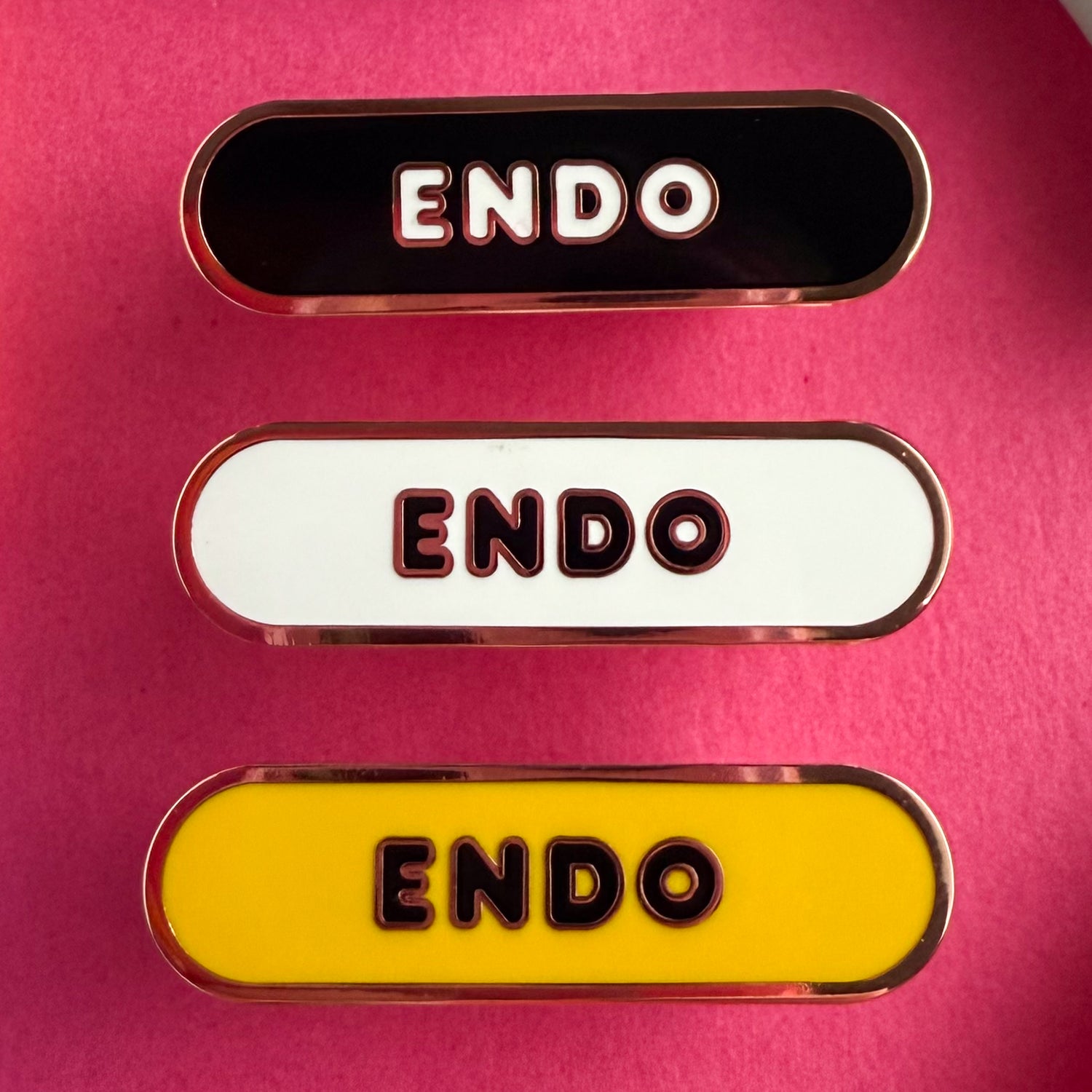 Three bandaid shaped pins with the text ENDO in bubble letters. Top one black with white text, the next is white with black text, and the last is yellow with black text. The pins are on a hot pink background.