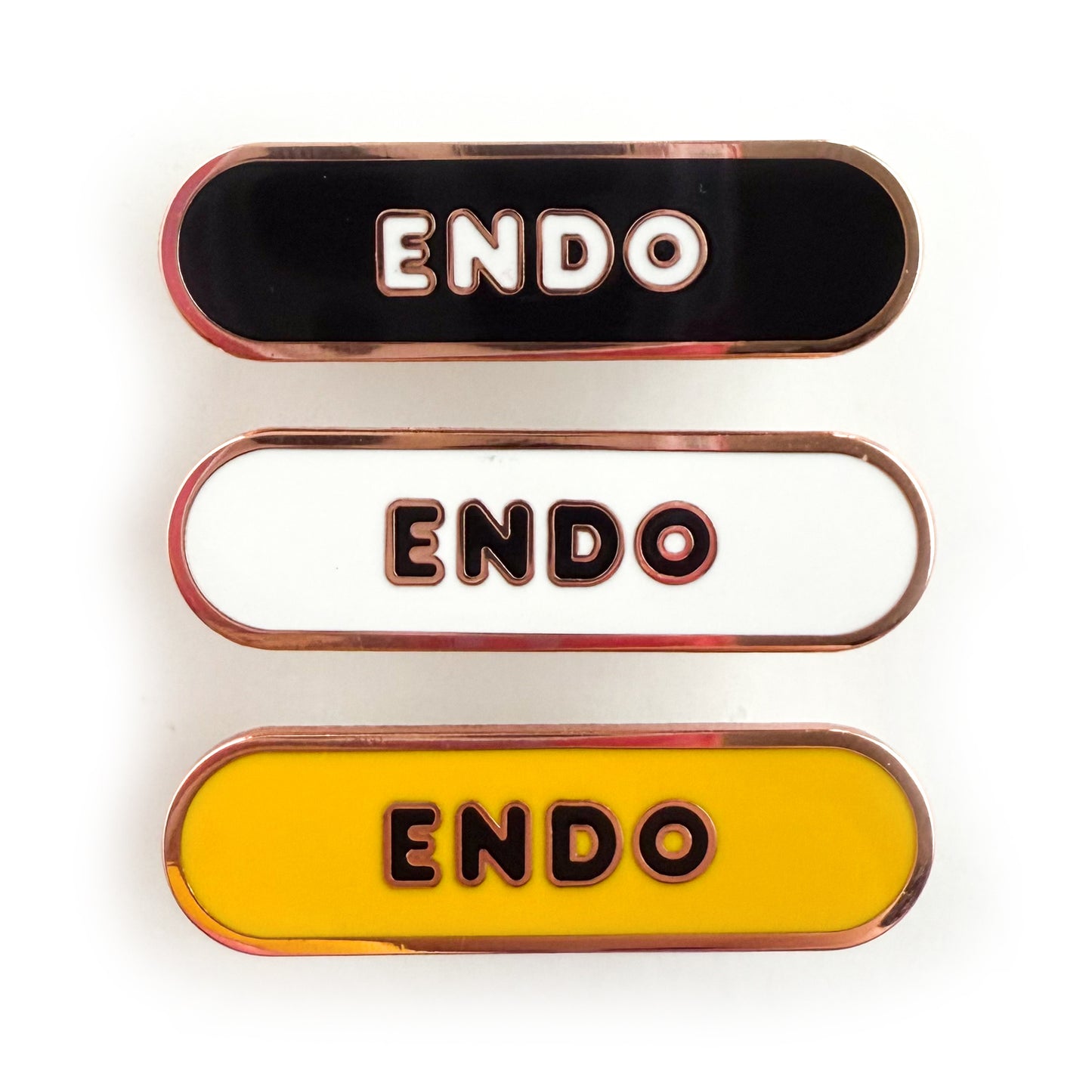 Three bandaid shaped pins with the text ENDO in bubble letters. Top one black with white text, the next is white with black text, and the last is yellow with black text.