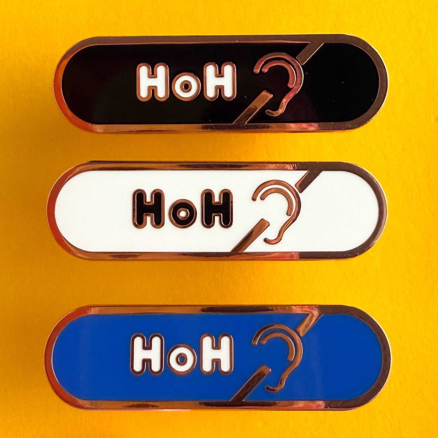 Three bandaid shaped pins with the text HoH in bubble letters and the hard of hearing symbol. Top one black with white text, the next is white with black text, and the last is blue with white text. The pins are on a yellow background.