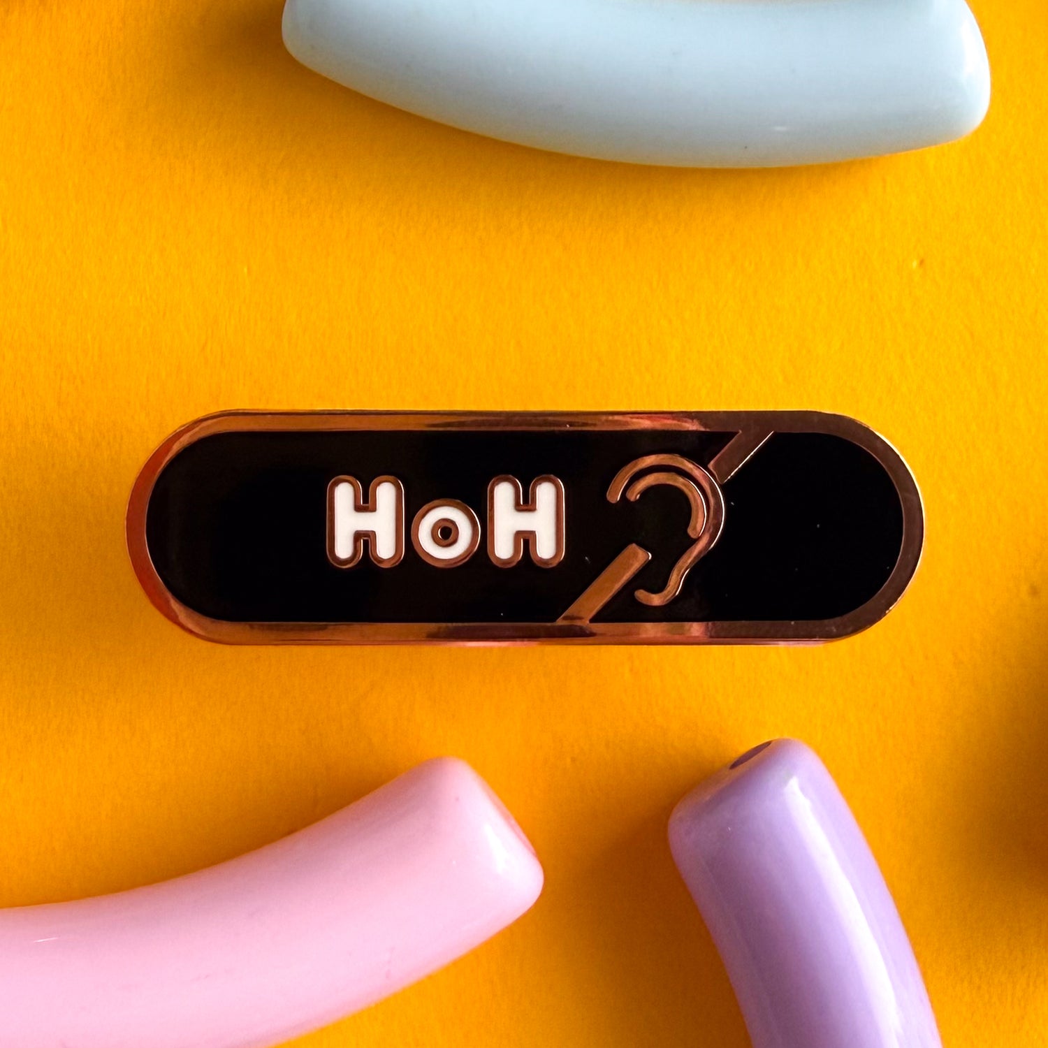 A bandaid shaped pin with the text HoH in bubble letters and the hard of hearing symbol. The background is black and the text is white. The pin is on a yellow background.