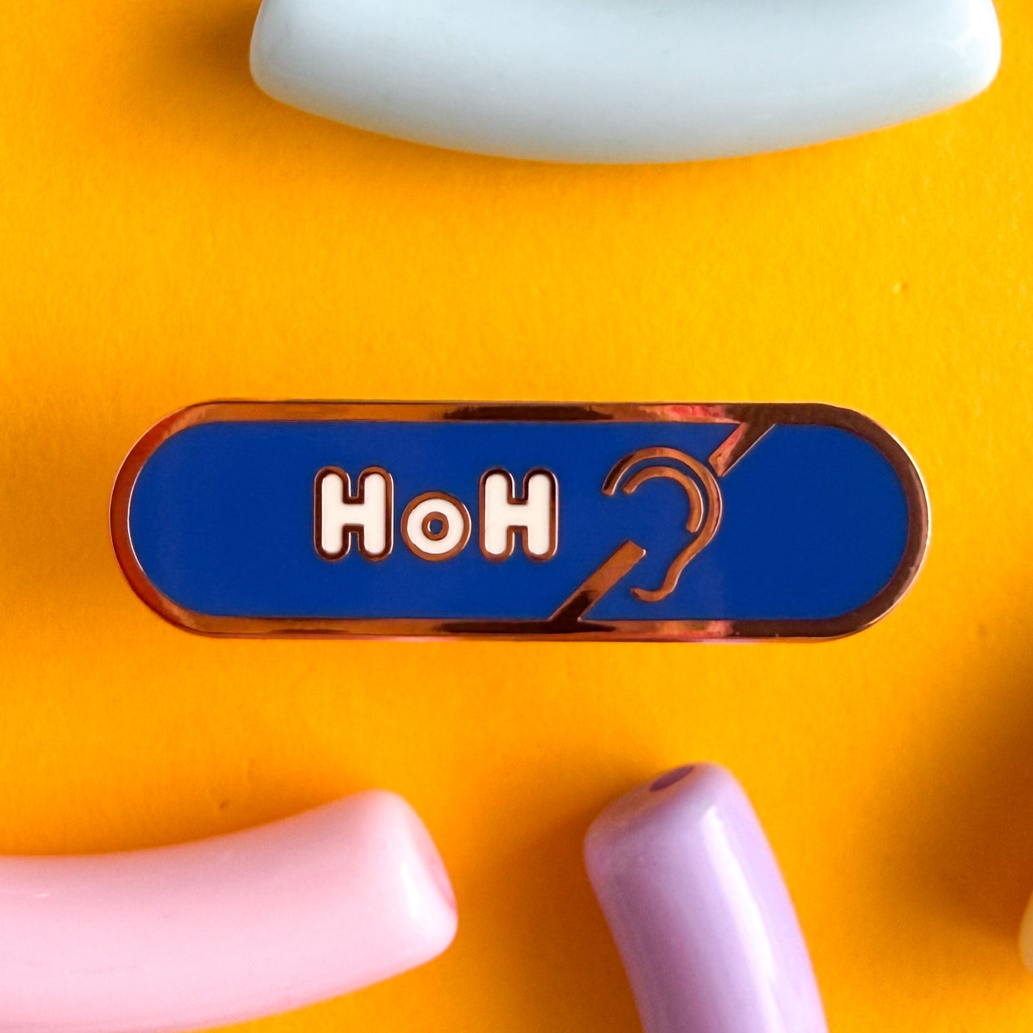 A bandaid shaped pin with the text HoH in bubble letters and the hard of hearing symbol. The background is blue and the text is white. The pin is on yellow background.