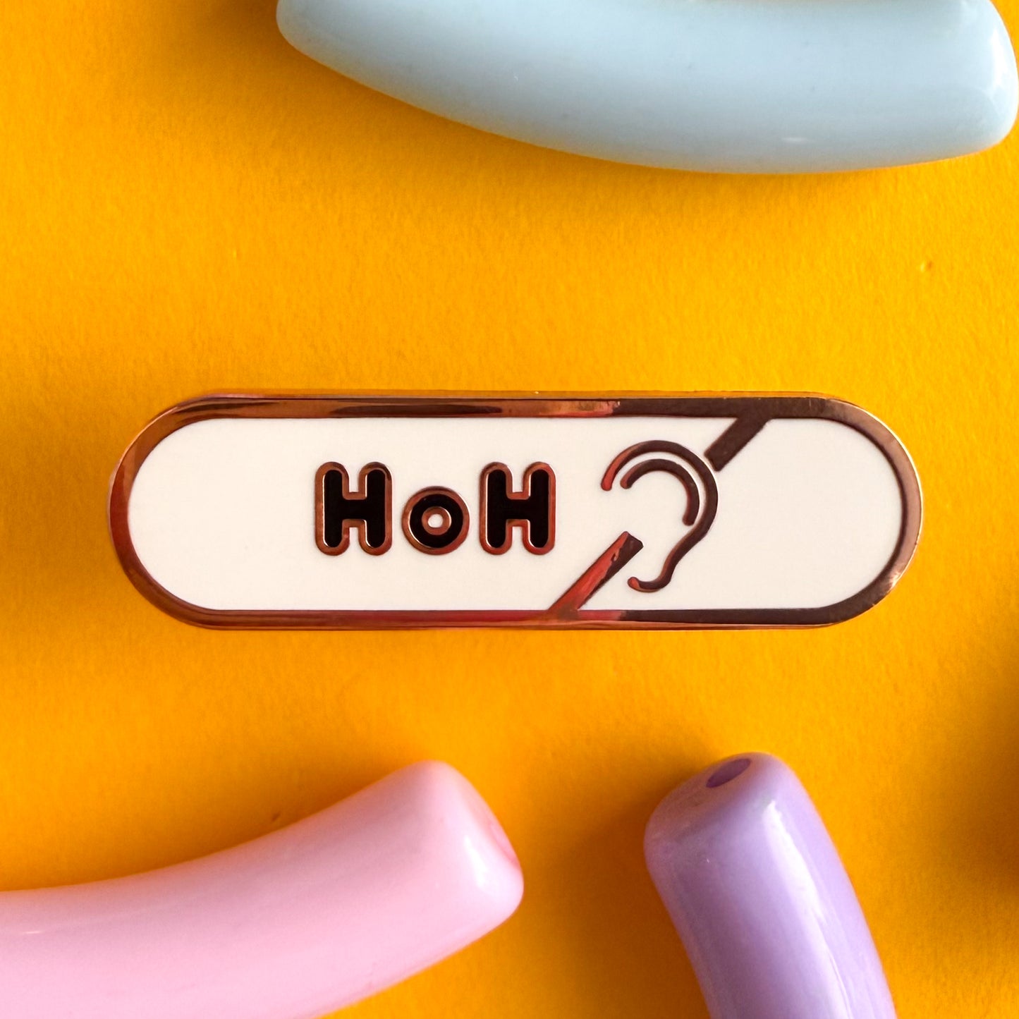 A bandaid shaped pin with the text HoH in bubble letters and the hard of hearing symbol. The background is white and the text is black. The pin is on a yellow background.