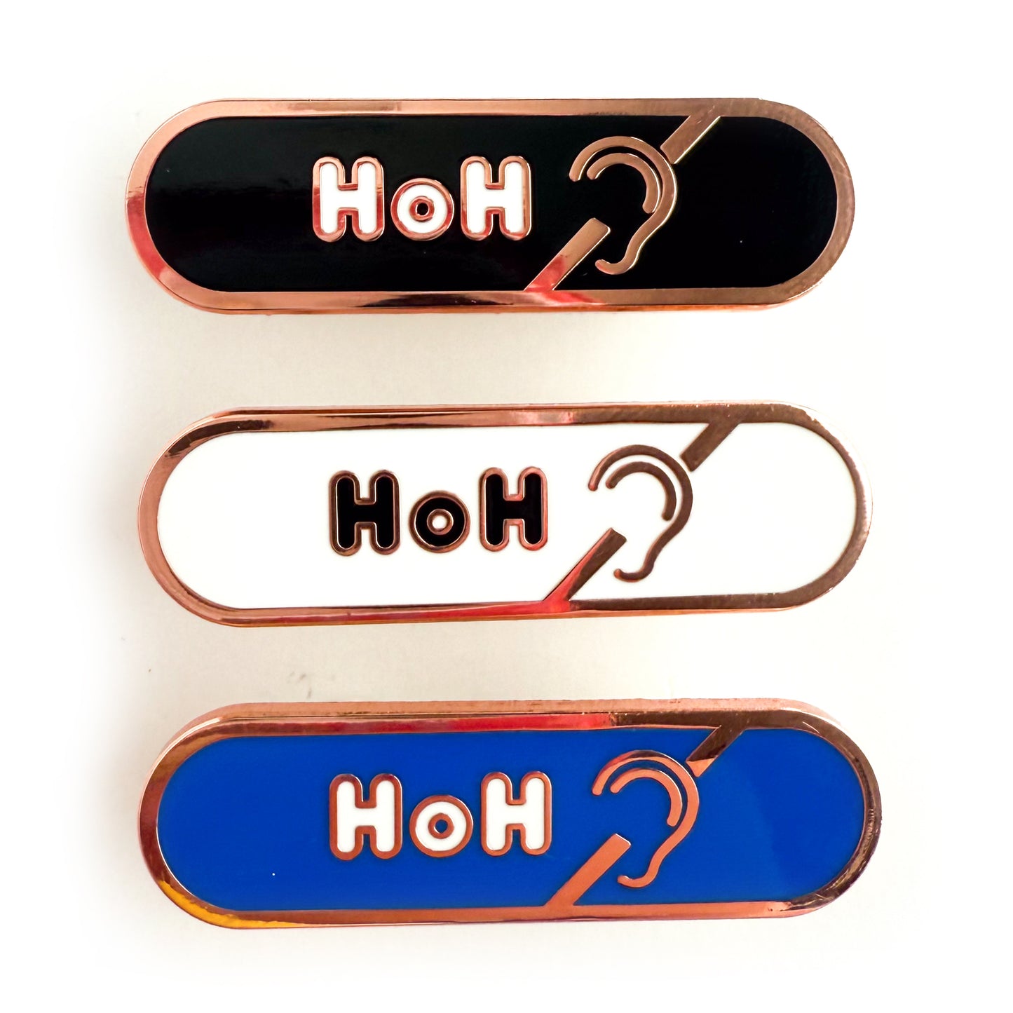 Three bandaid shaped pins with the text HoH in bubble letters and the hard of hearing symbol. Top one black with white text, the next is white with black text, and the last is blue with white text.