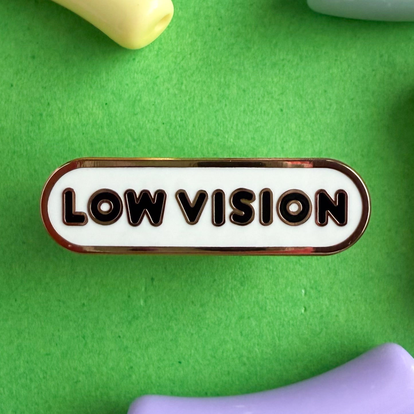 A bandaid shaped pin with the text LOW VISION  in bubble letters. The background is white and the text is black. The pin is on a green background.
