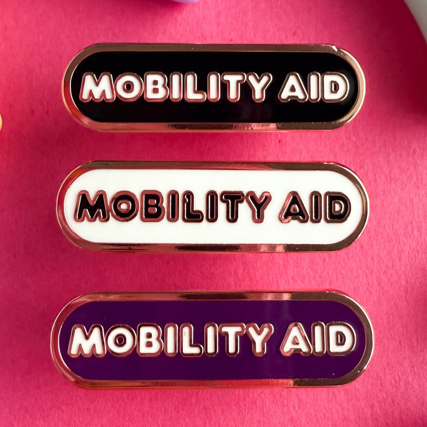 Three bandaid shaped pins with the text MOBILITY AID in bubble letters. Top one black with white text, the next is white with black text, and the last is eggplant purple with white text. The pins are on a pink background.