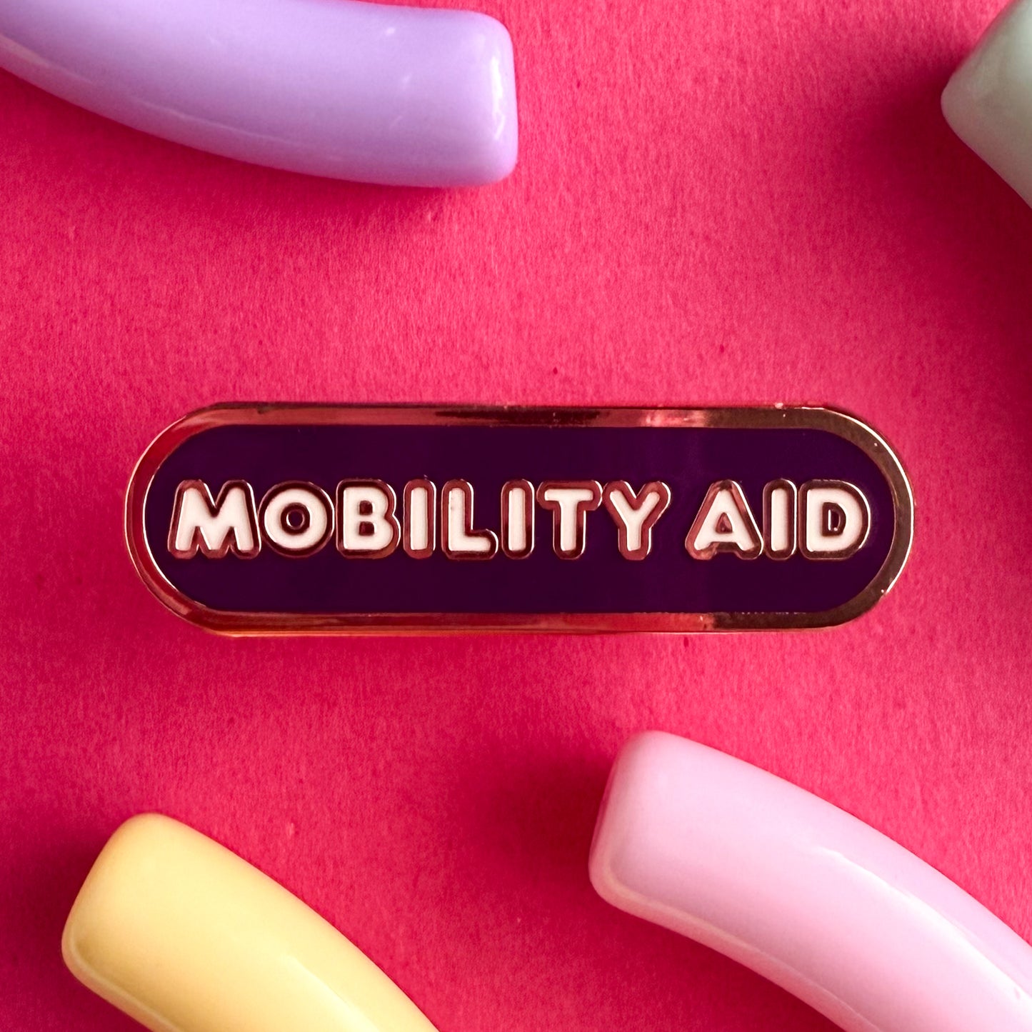 A bandaid shaped pin with the text MOBILITY AID in bubble letters. The background is dark purple and the text is white. The pin is on a pink background.