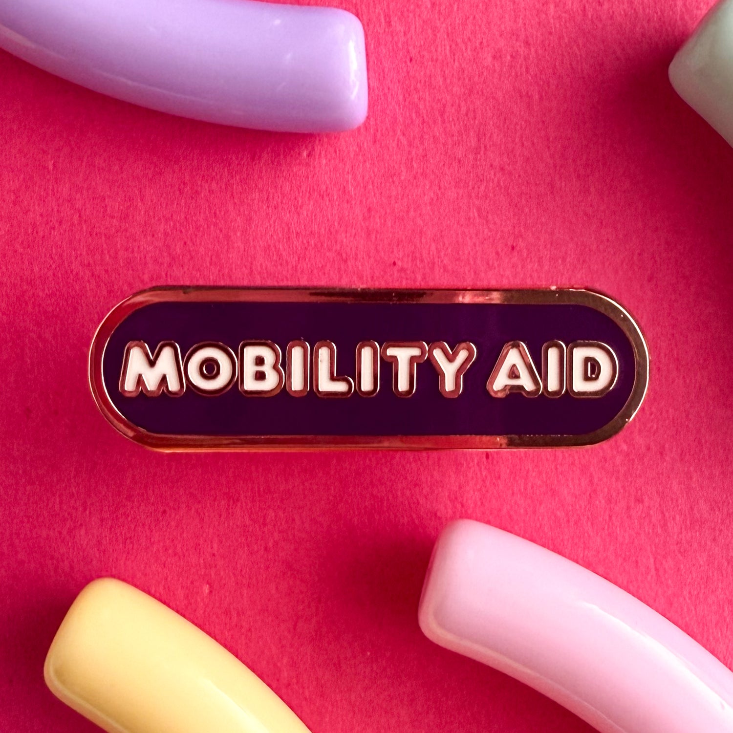 A bandaid shaped pin with the text MOBILITY AID in bubble letters. The background is dark purple and the text is white. The pin is on a pink background.