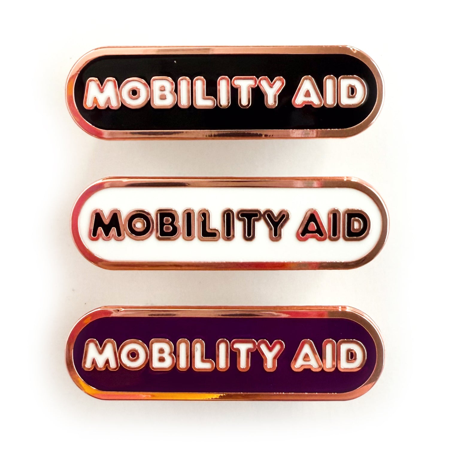 Three bandaid shaped pins with the text MOBILITY AID in bubble letters. Top one black with white text, the next is white with black text, and the last is eggplant purple with white text.