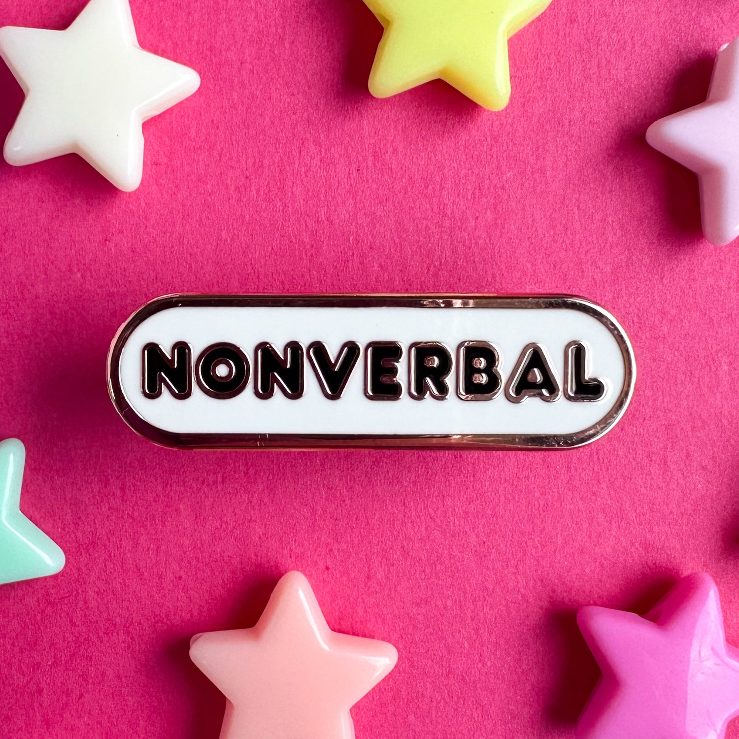 A white oval shaped enamel pin that reads "Nonverbal". The pin is sitting on a hot pink background with plastic star beads around it.