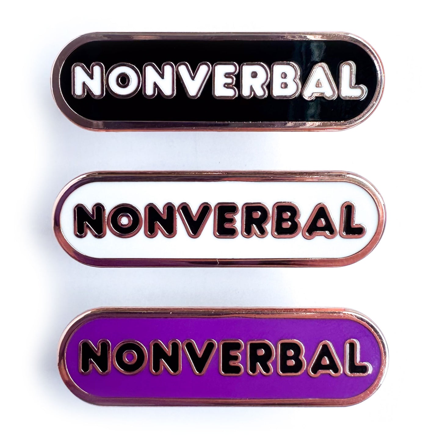 Three capsule shaped enamel pins that all have bubble letters that read "Nonverbal", the top pin is black, the middle is white, and the bottom is purple.