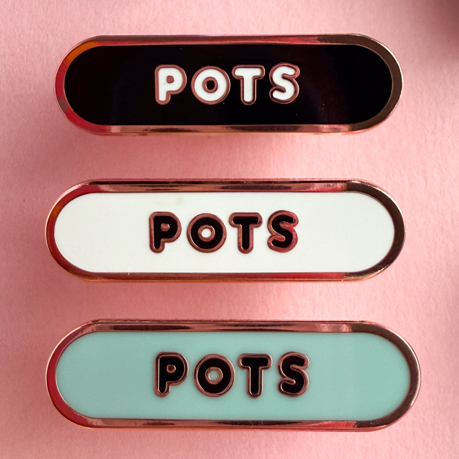 Three bandaid shaped pin with the text POTS in bubble letters. Top one black with white text, the next is white with black text, and the last is light blue with black text. The pins are on a pink background.