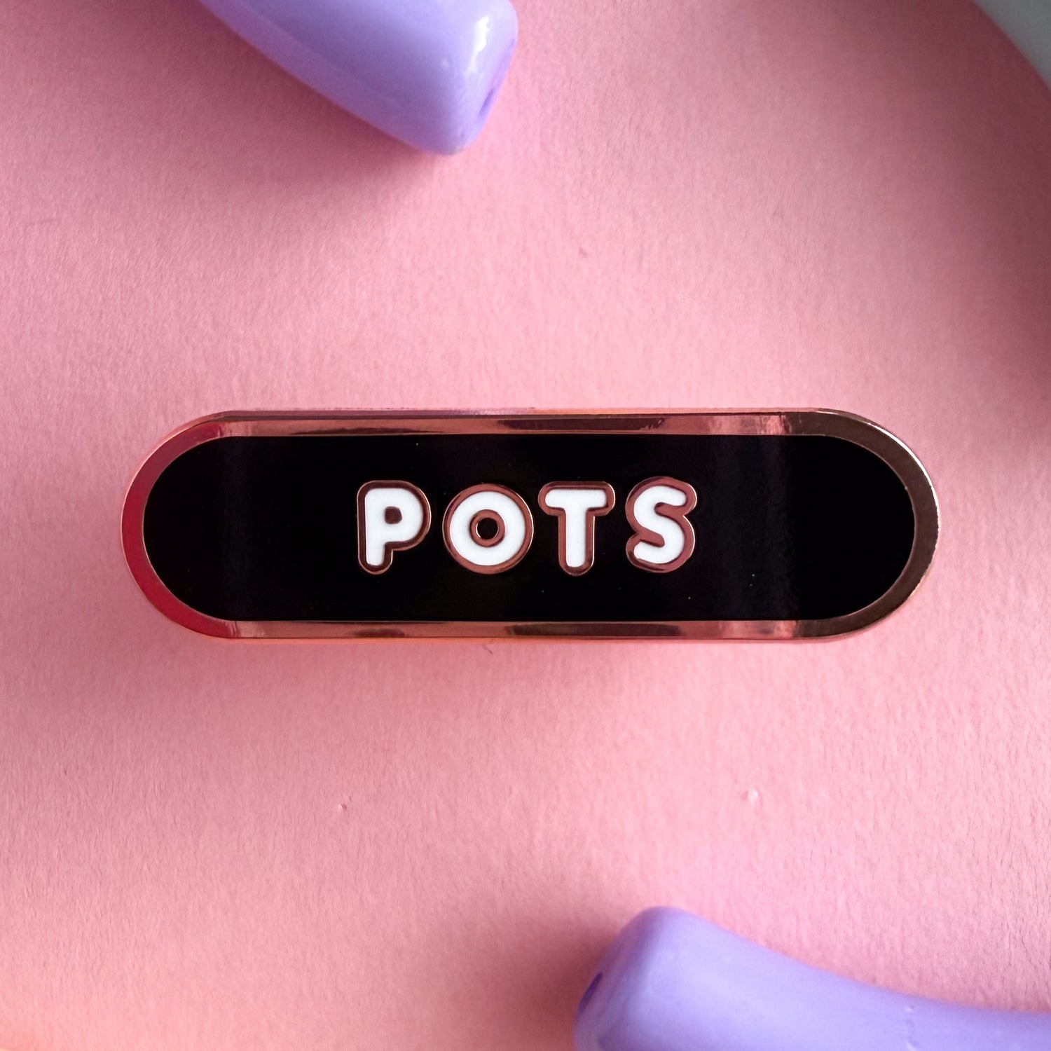 A bandaid shaped pin with the text POTS in bubble letters. The background is black and the text is white. The pin is on a purple background.