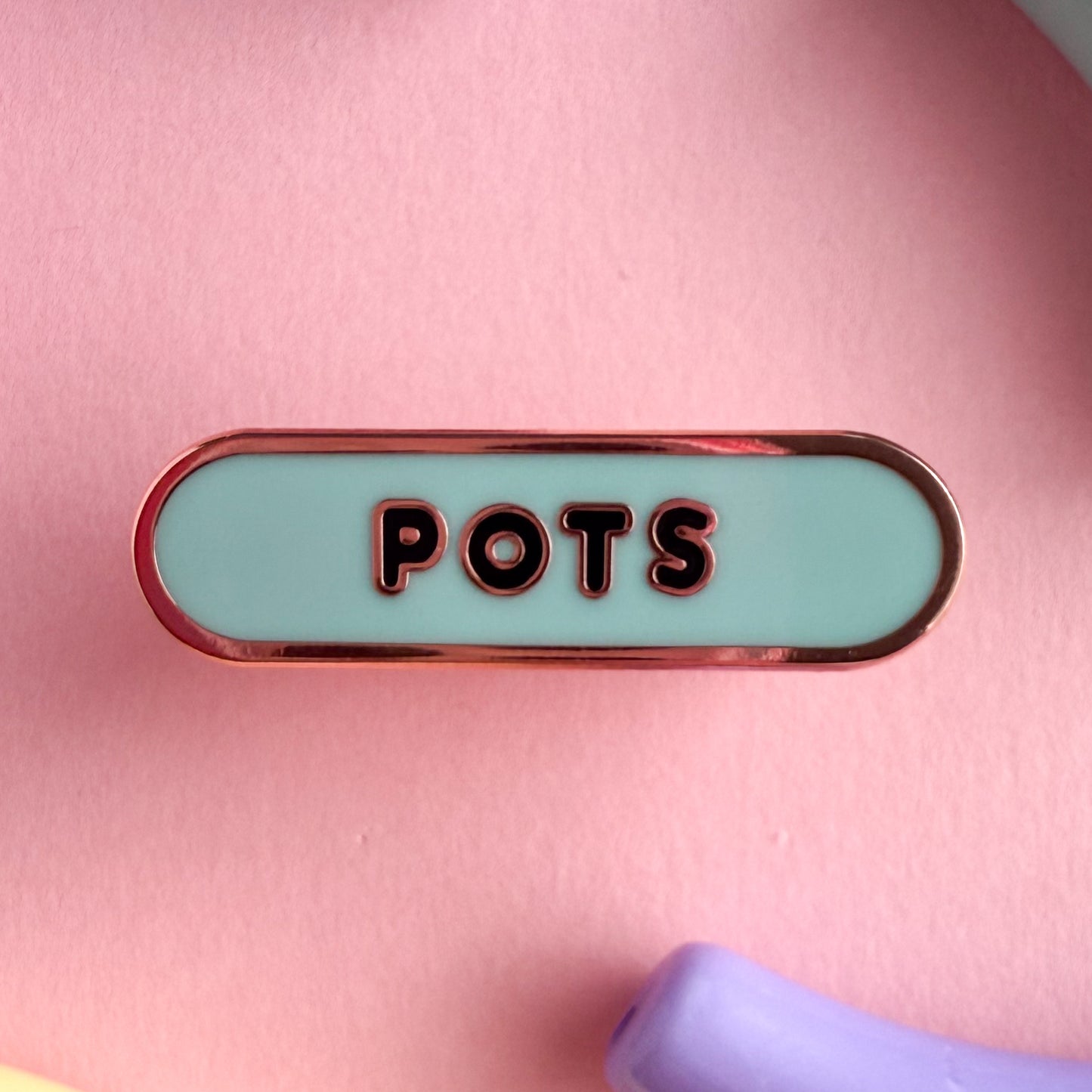 A bandaid shaped pin with the text POTS in bubble letters. The background is light blue and the text is black. The pin is on a pink background.