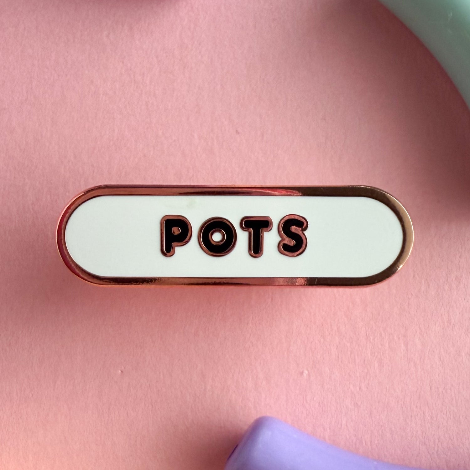 A bandaid shaped pin with the text POTS in bubble letters. The background is white and the text is black. The pin is on a pink background.