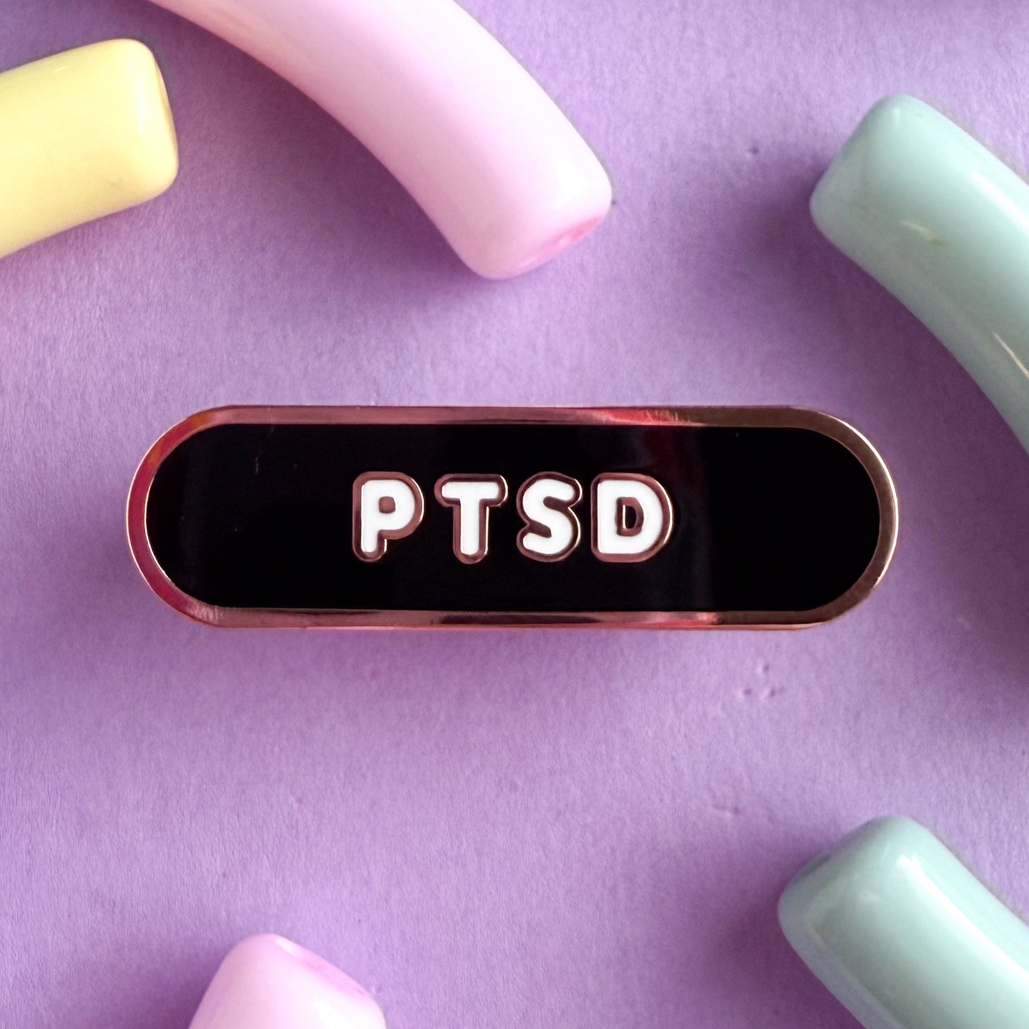 Abandaid shaped pin with the text PTSD in bubble letters. The background is black and the text is white. The pin is on a purple background.
